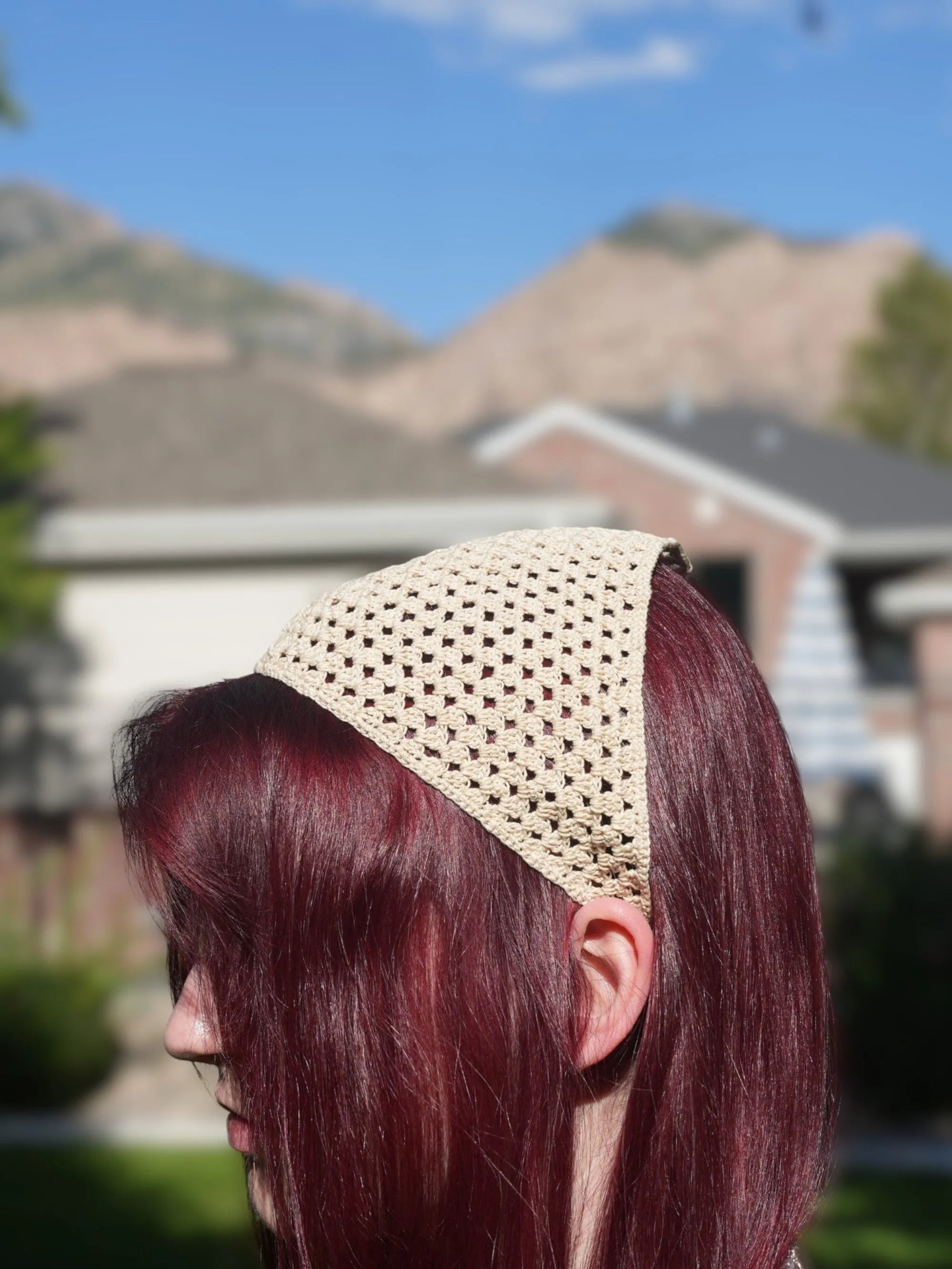 Person with long, red hair wearing a beige crochet headscarf, pictured outdoors with blurred mountains and houses in the background.