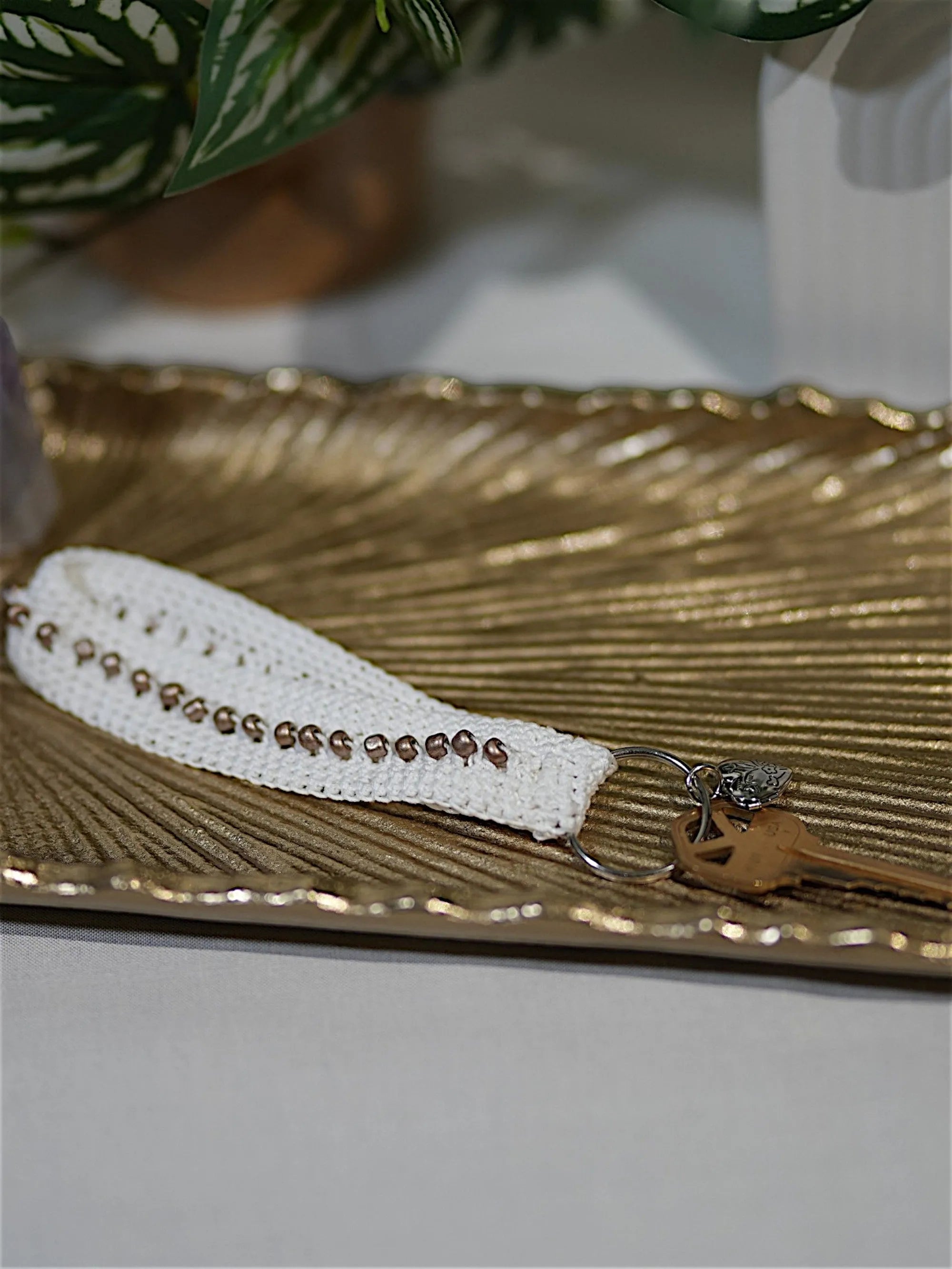 A stylish, handmade keychain with white crochet and bead details, resting on an elegant gold decorative tray.