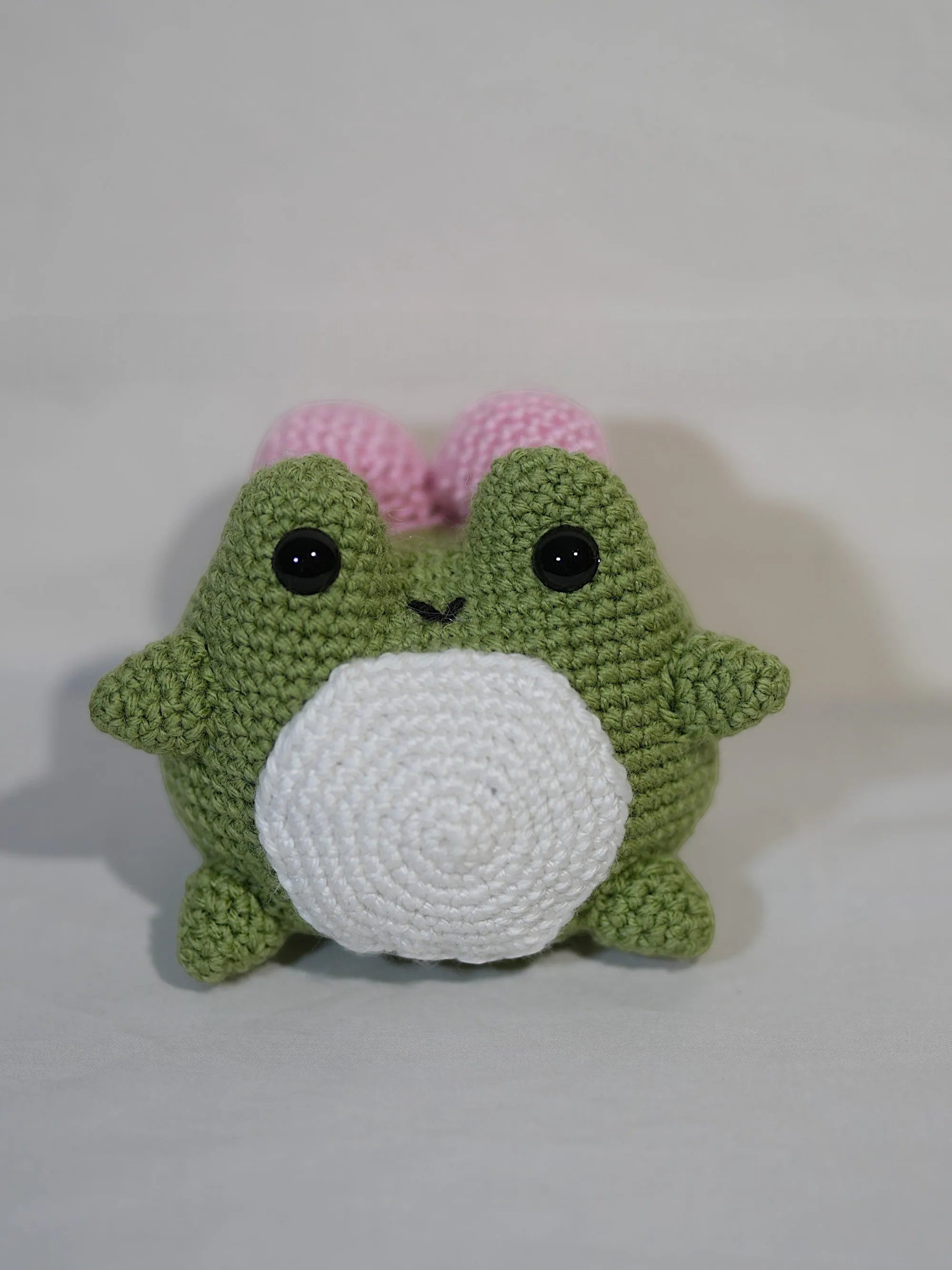 A handmade green amigurumi frog with black eyes and a white belly, featuring pink cheeks, set against a plain background.