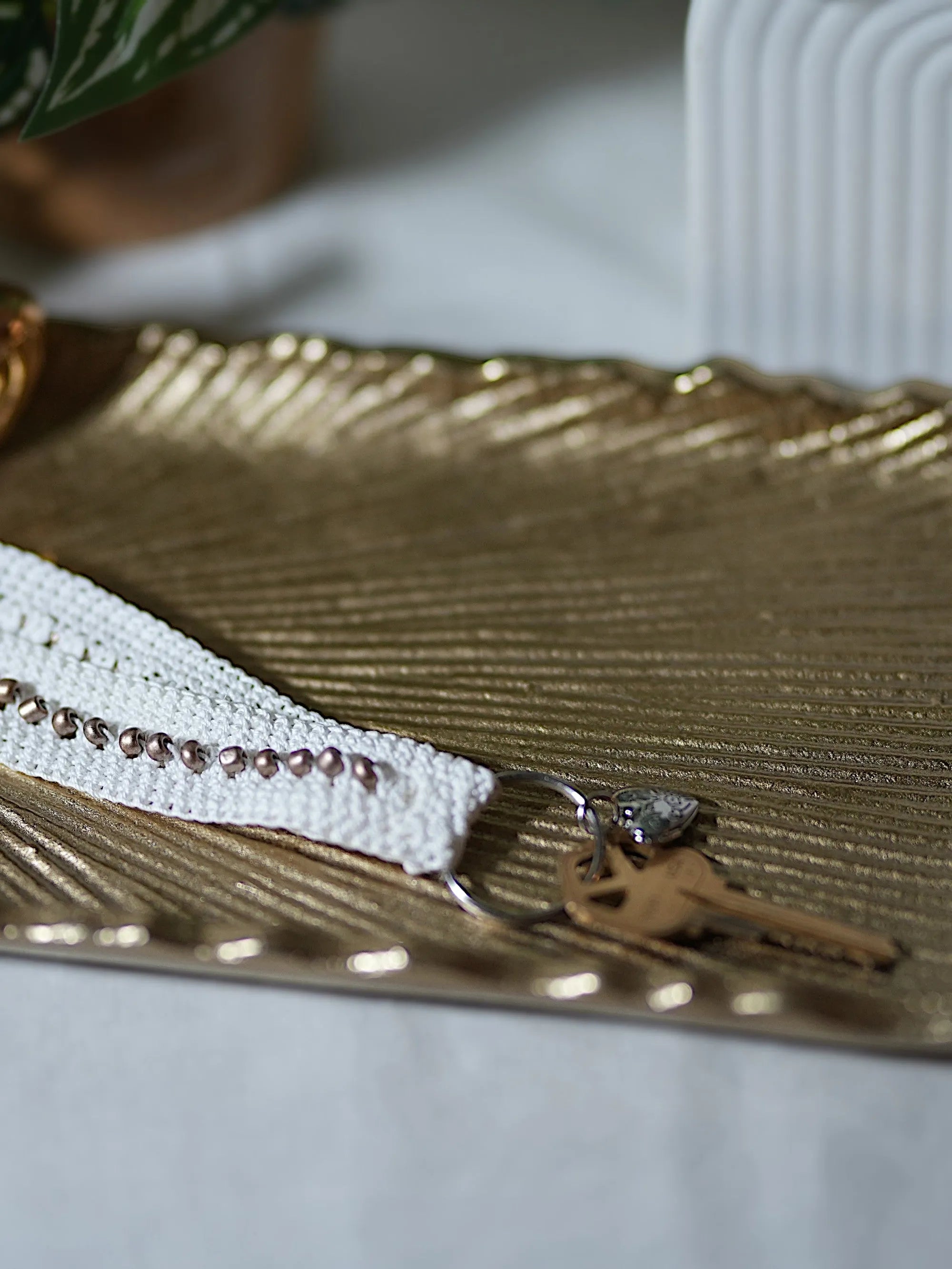 A handcrafted beaded keychain with a white crocheted band and metallic beads rests on a textured gold tray.