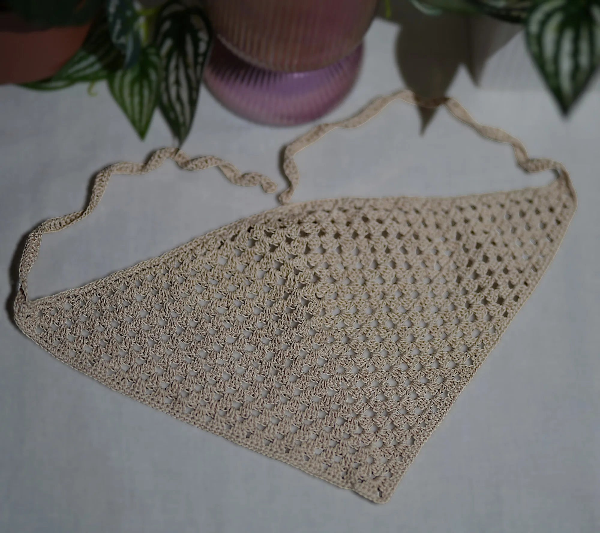 A beige crocheted triangular scarf with intricate patterns spread out on a table beneath a pink vase and green leaves.