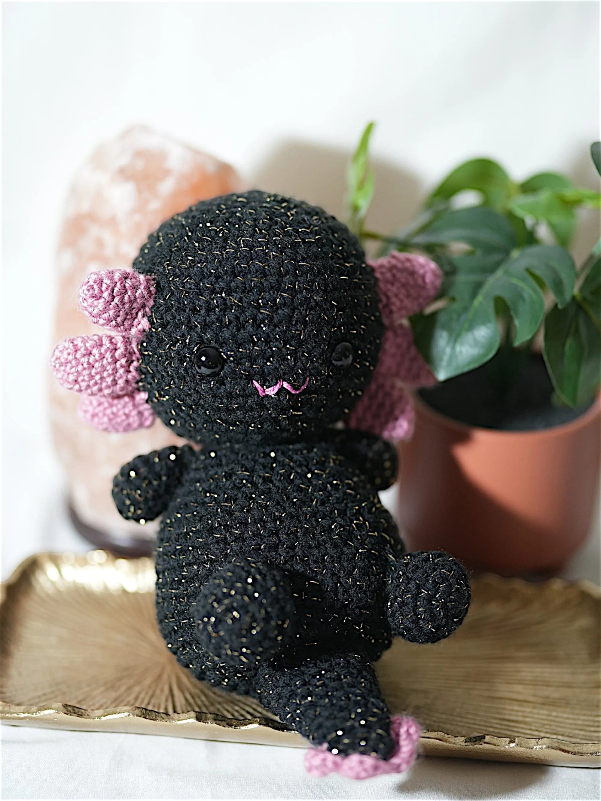 A black crocheted axolotl plush toy with pink accents sits on a decorative tray, surrounded by a pink salt lamp and a potted plant.