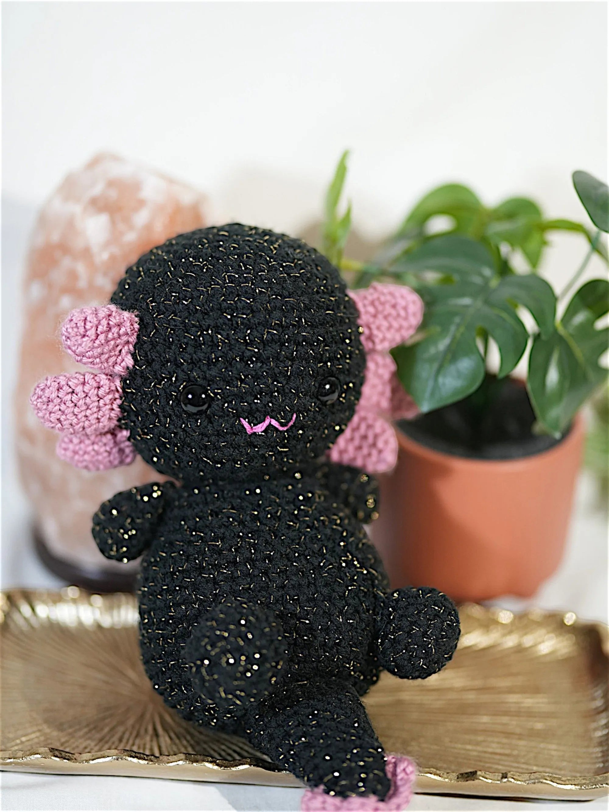 A black crocheted axolotl plush toy with pink accents sits on a decorative tray, surrounded by a pink salt lamp and a potted plant.