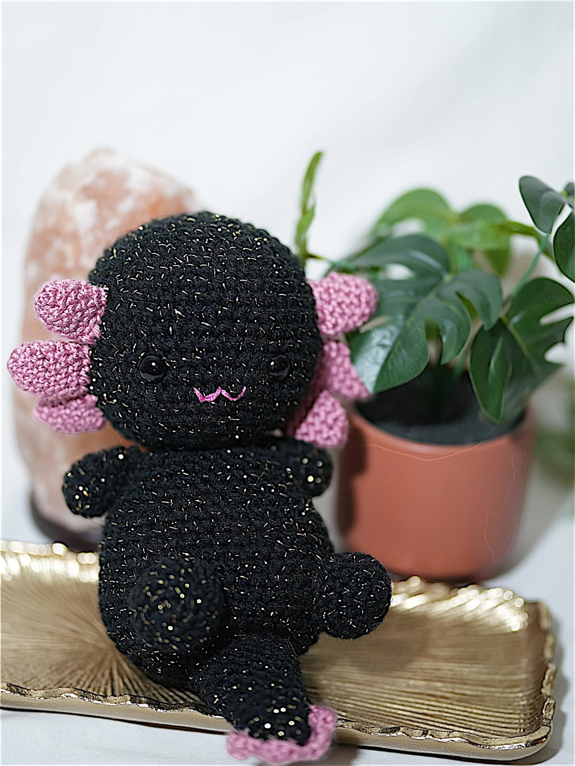 A cute handmade black crocheted axolotl toy with pink accents, resting on a decorative tray with plants in the background.