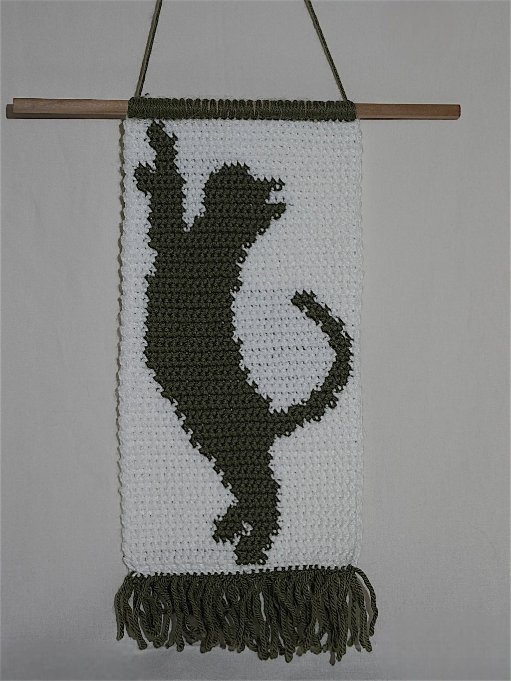 A woven wall hanging depicting a silhouette of a playful cat in dark yarn on a white background, featuring fringe at the bottom and mounted on a wooden rod.