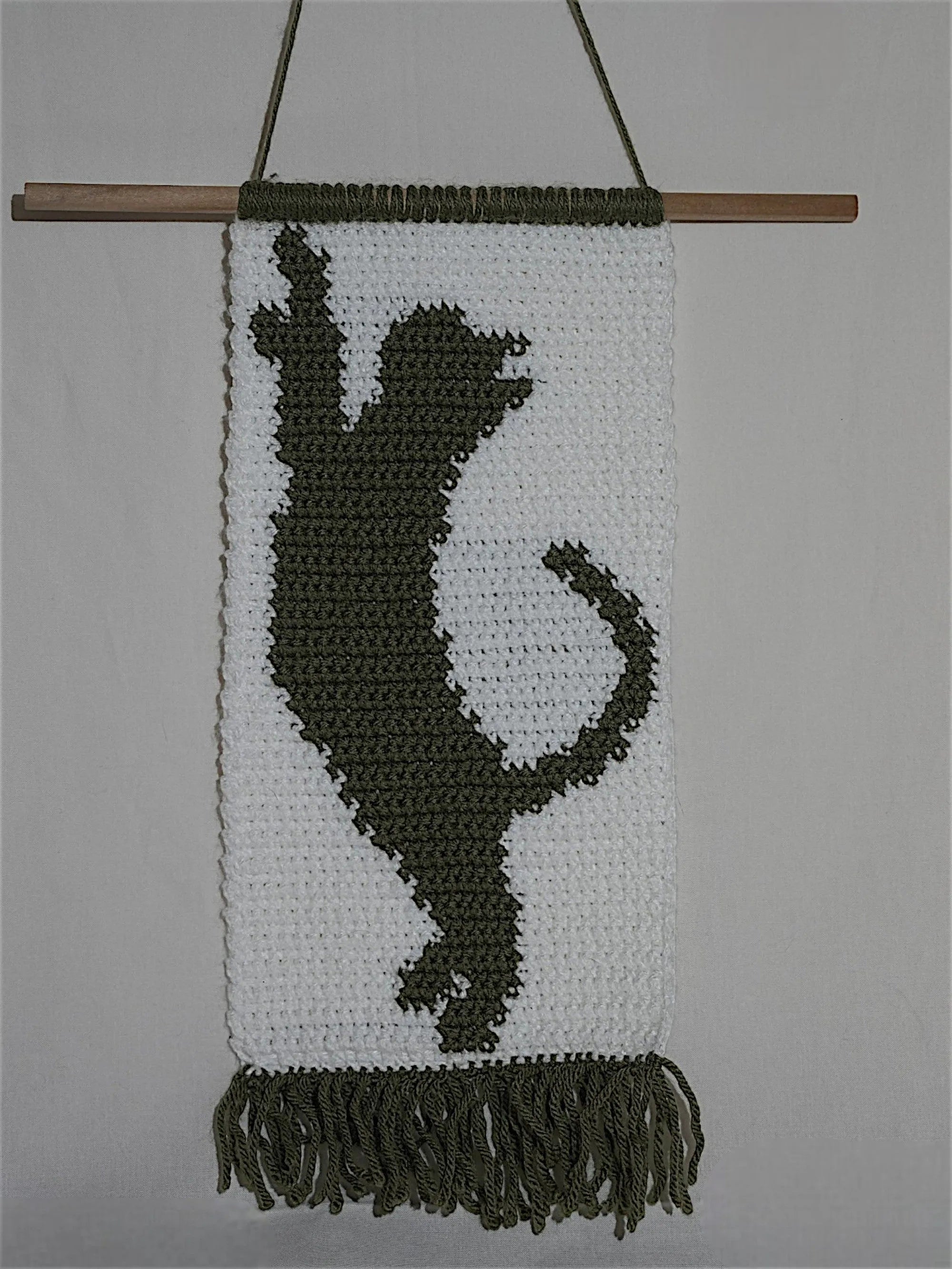 A woven wall hanging depicting a silhouette of a playful cat in dark yarn on a white background, featuring fringe at the bottom and mounted on a wooden rod.