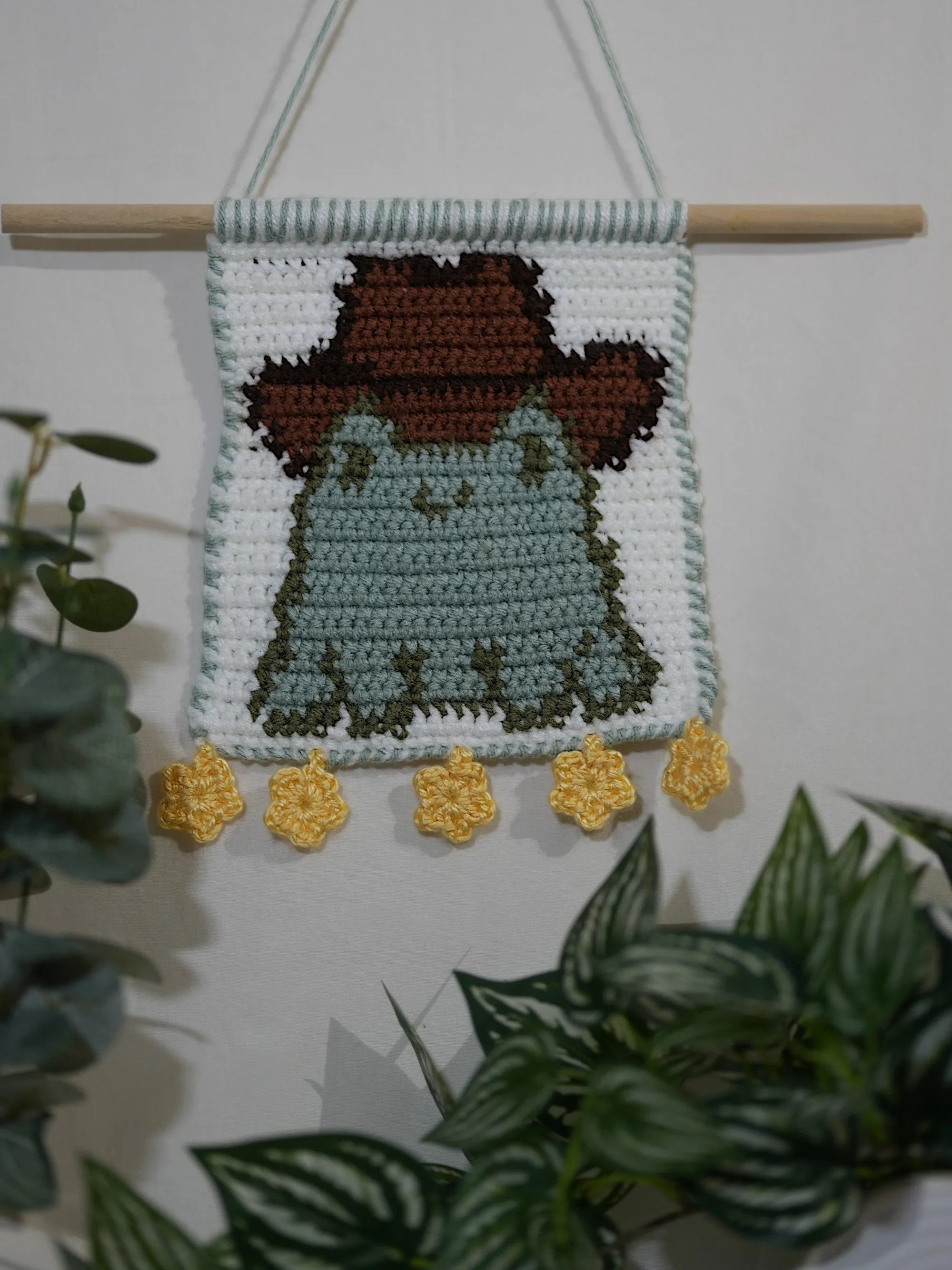 A crochet wall hanging featuring a frog wearing a brown cowboy hat, displayed on a wooden dowel with yellow flower embellishments at the bottom, surrounded by green plants.