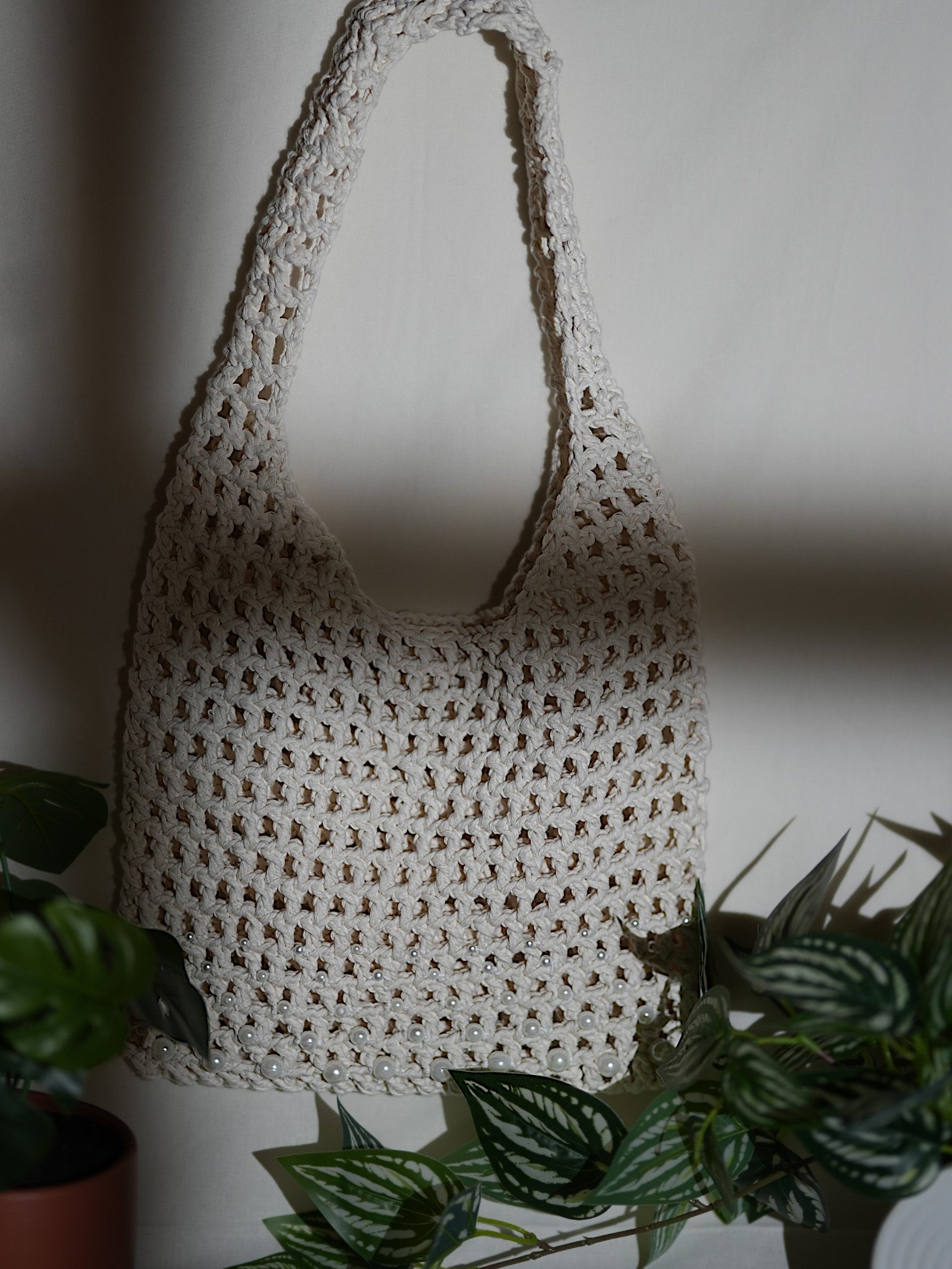 A handmade white crochet bag with an intricate lattice pattern, displayed against a plain background surrounded by green plants.