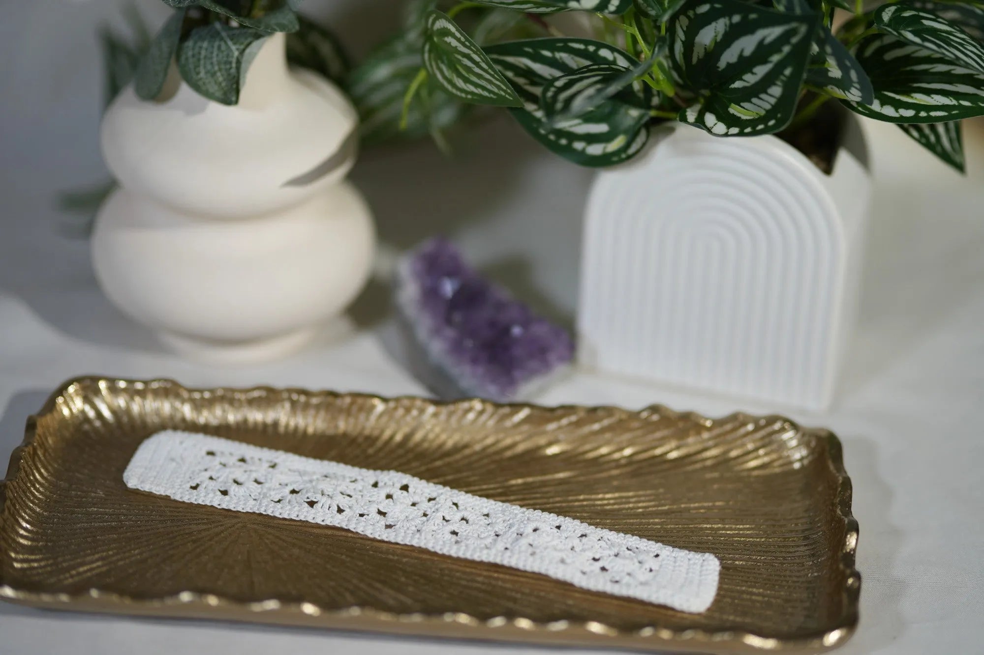 A decorative white crochet bookmark on a textured golden tray, accompanied by a potted plant and a small purple crystal.