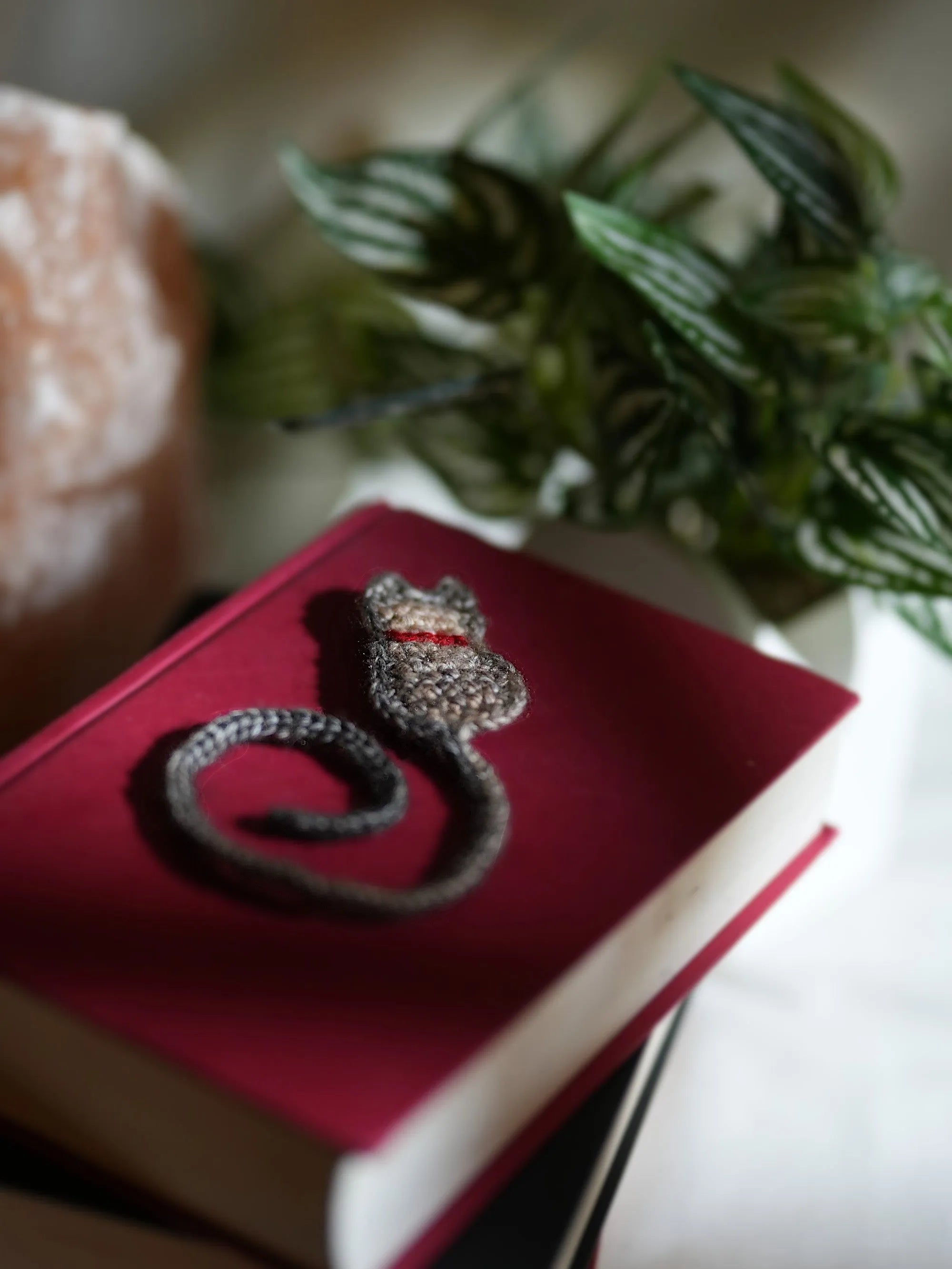 A handmade crochet cat bookmark resting on a red hardcover book beside a plant.