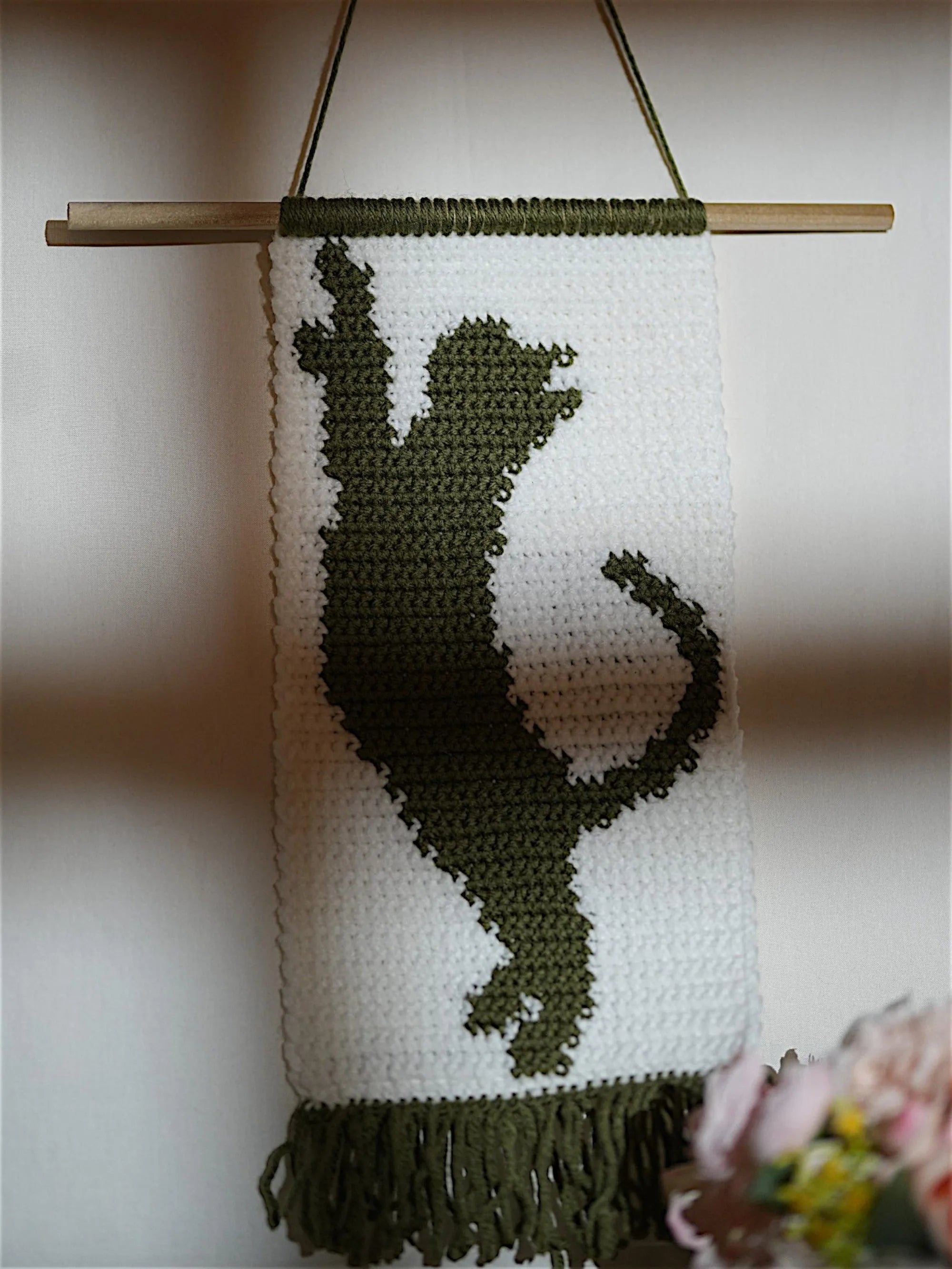 A crocheted wall hanging with a green silhouette of a playful cat on a white background, finished with green fringe.