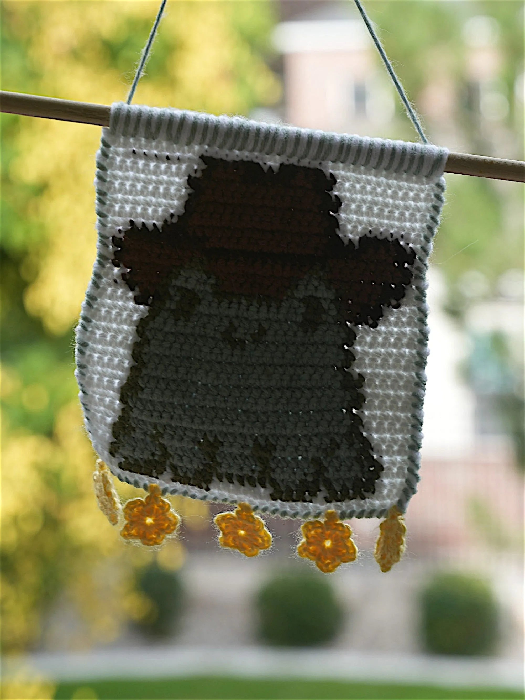 A crocheted wall hanging featuring a cute character with a brown hat, embellished with yellow flower ornaments at the bottom.