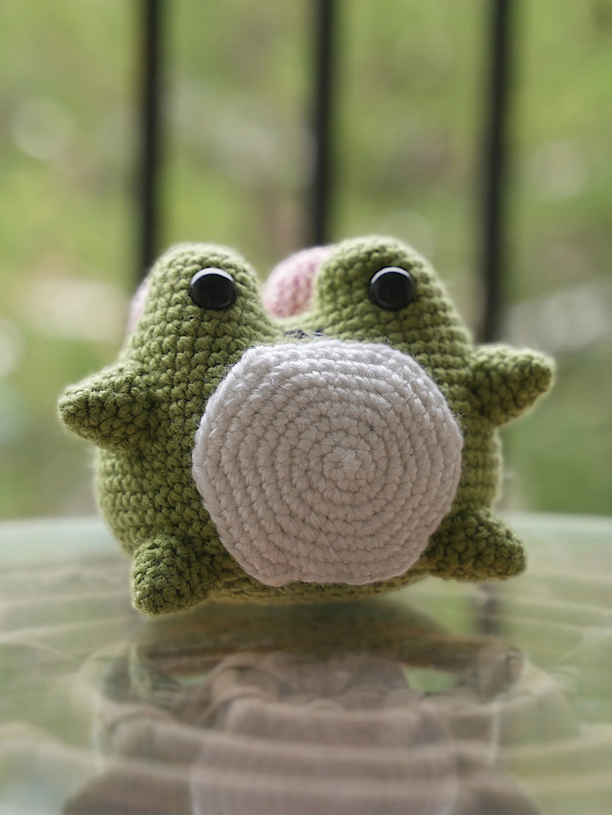 Handmade green crocheted dinosaur toy with a white belly, sitting on a reflective surface with a blurred background.