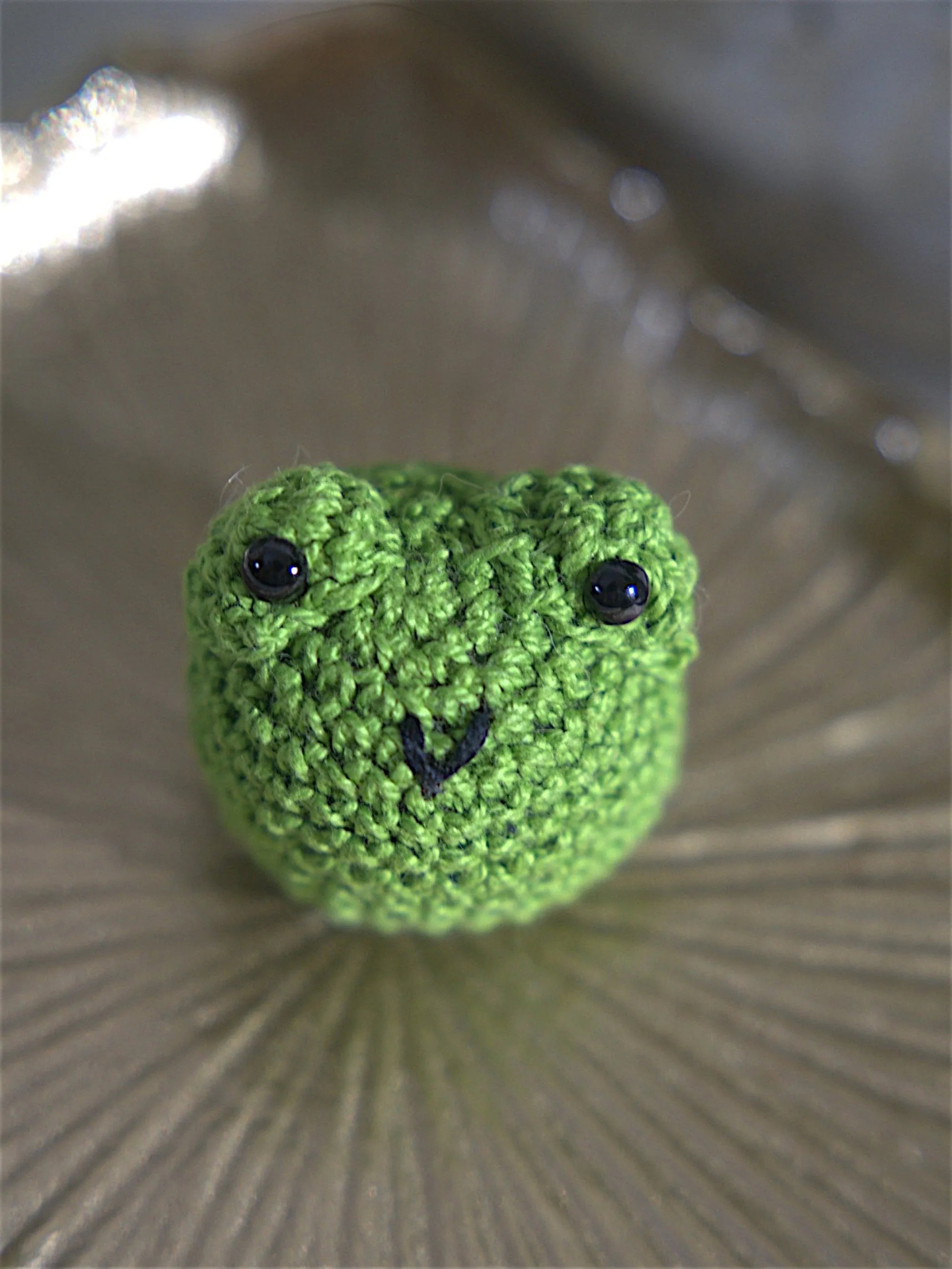 A handmade crochet green frog with black button eyes, sitting on a textured surface.