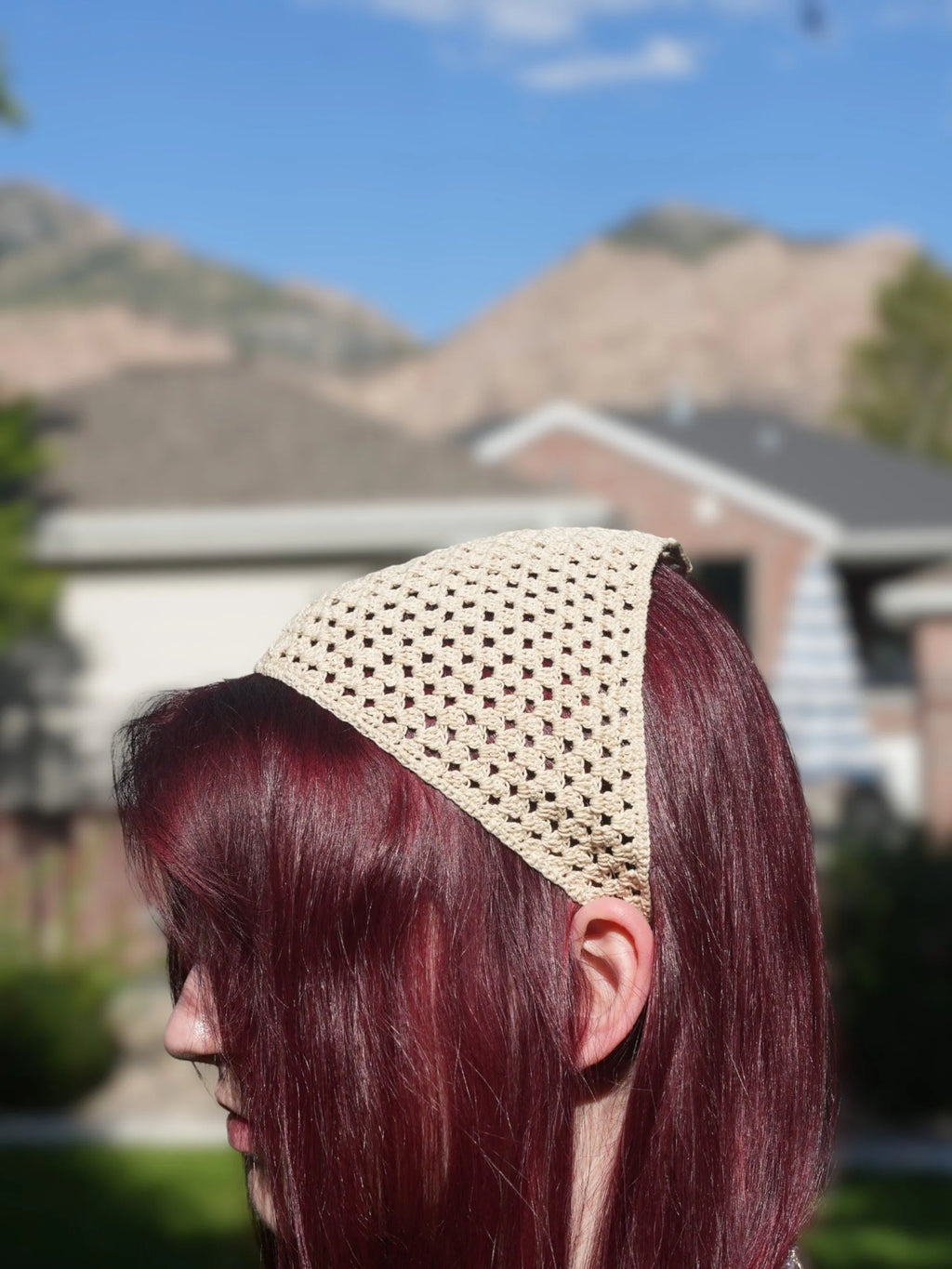 Person with long, red hair wearing a beige crochet headscarf, pictured outdoors with blurred mountains and houses in the background.