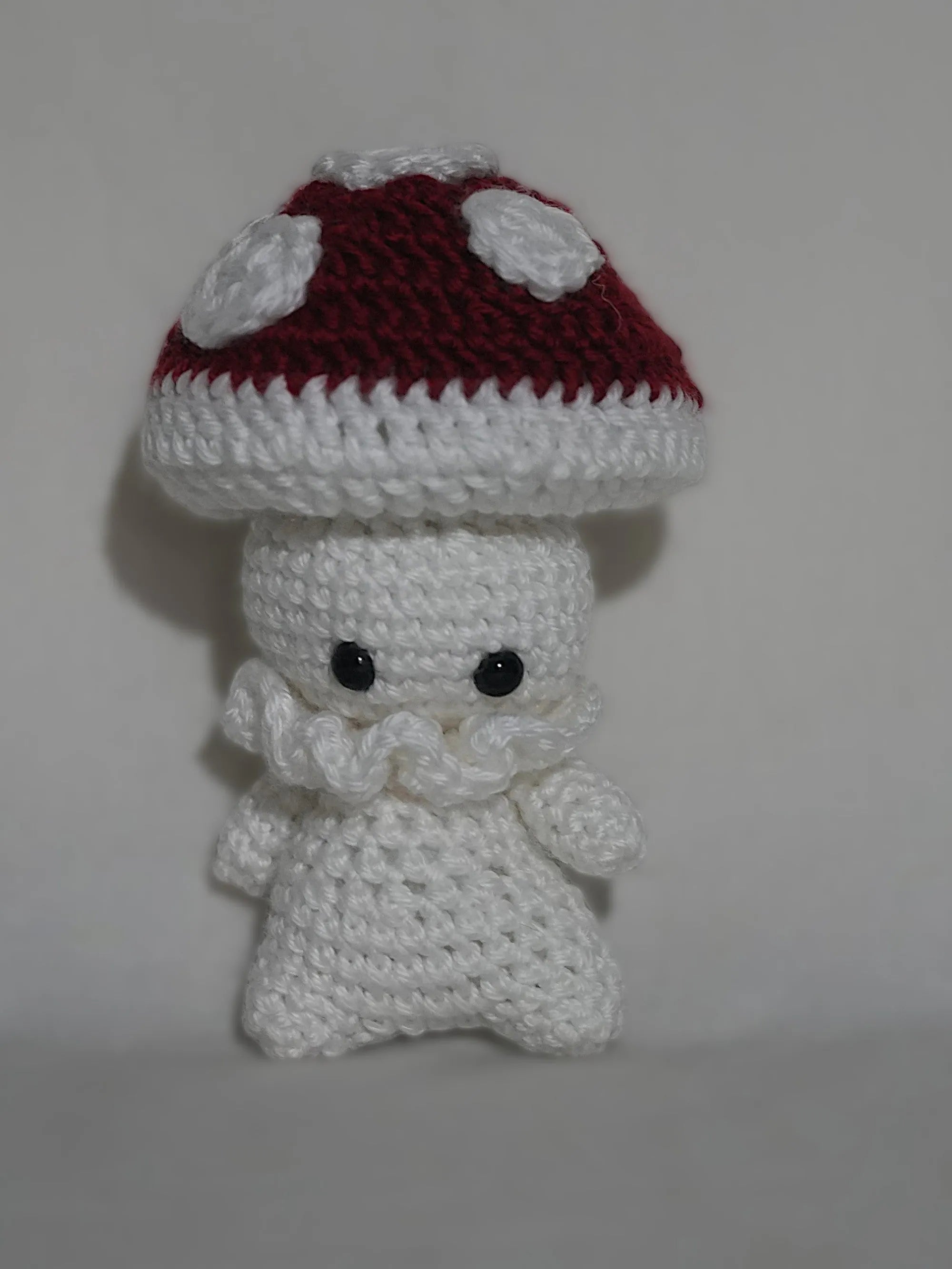 A handmade crochet toy resembling a mushroom character with a red and white cap and black eyes.