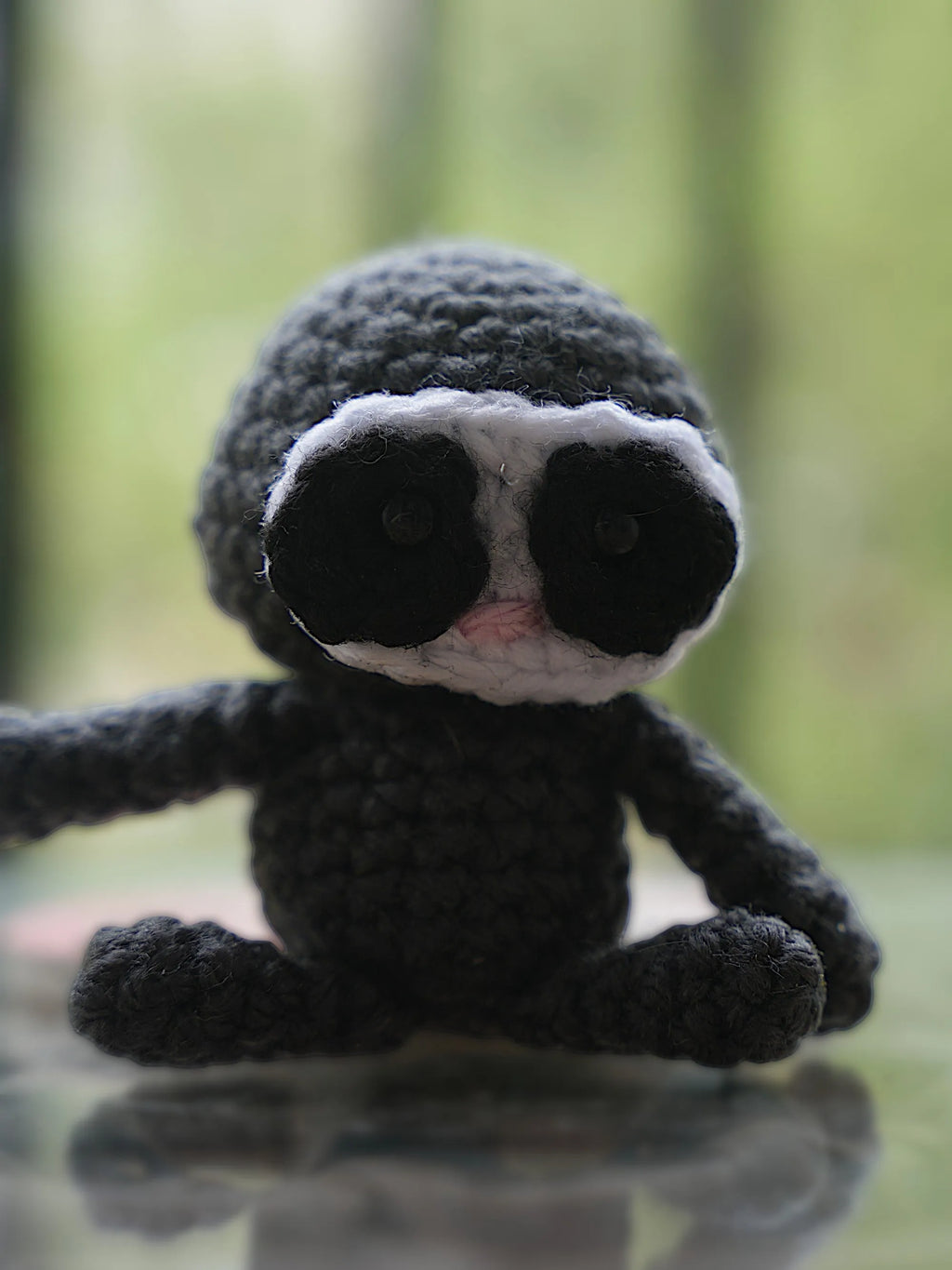 A small handmade crochet sloth toy sitting on a reflective surface with a blurred natural background.