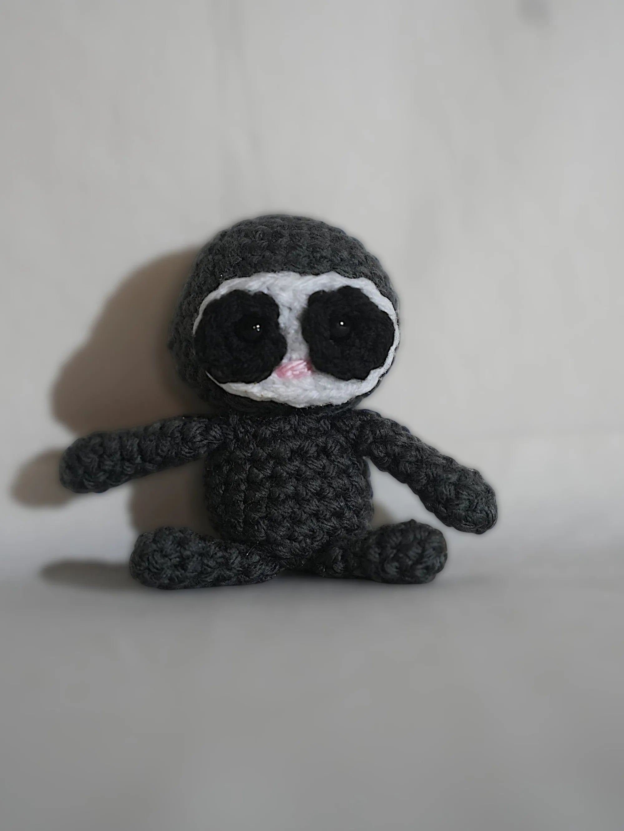 A handmade crochet sloth toy sitting upright with a blend of grey and white yarn on a plain background.