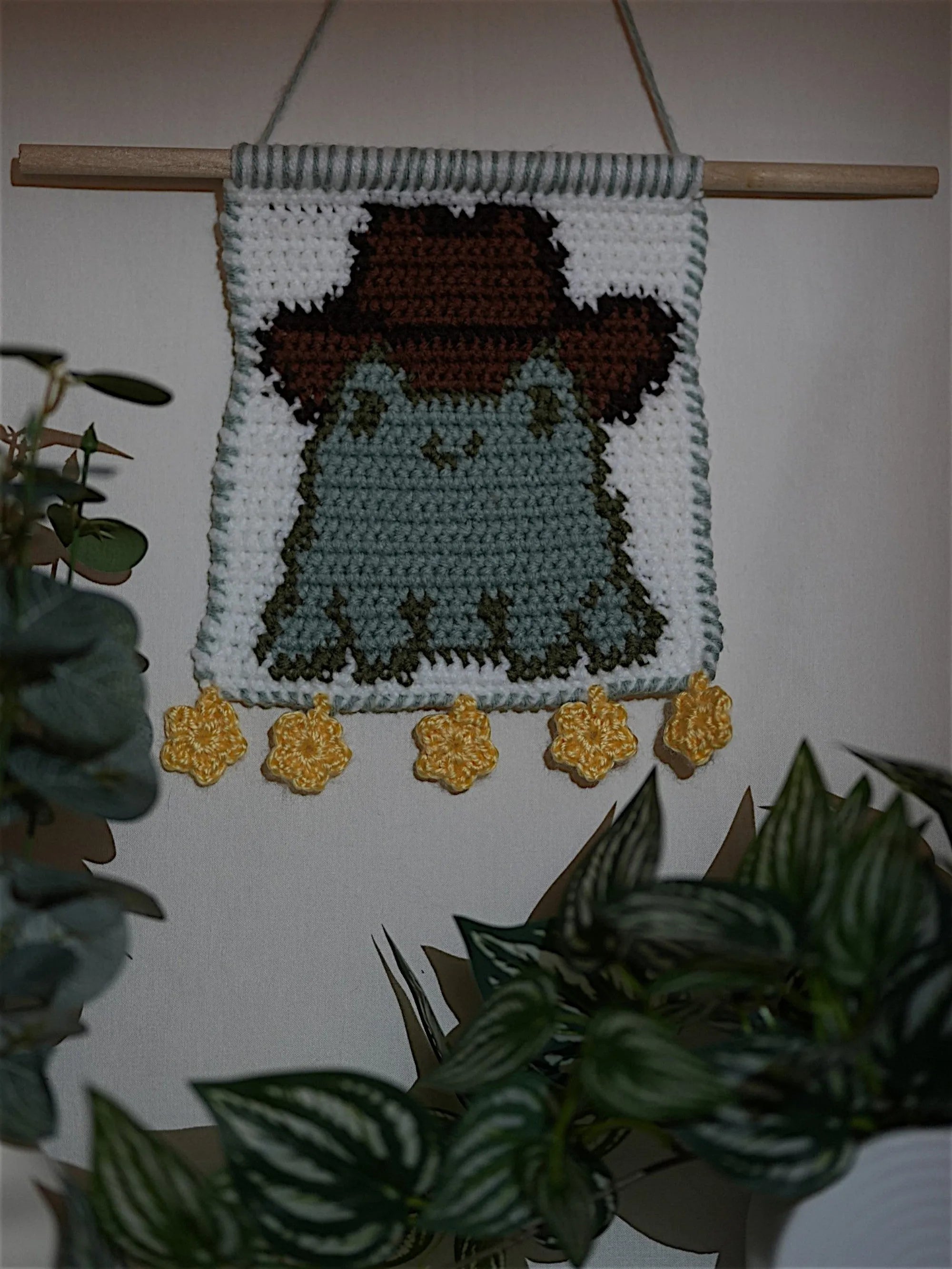 A crochet wall hanging featuring a green frog wearing a brown cowboy hat, displayed on a wooden dowel with yellow flower embellishments at the bottom, surrounded by green plants.