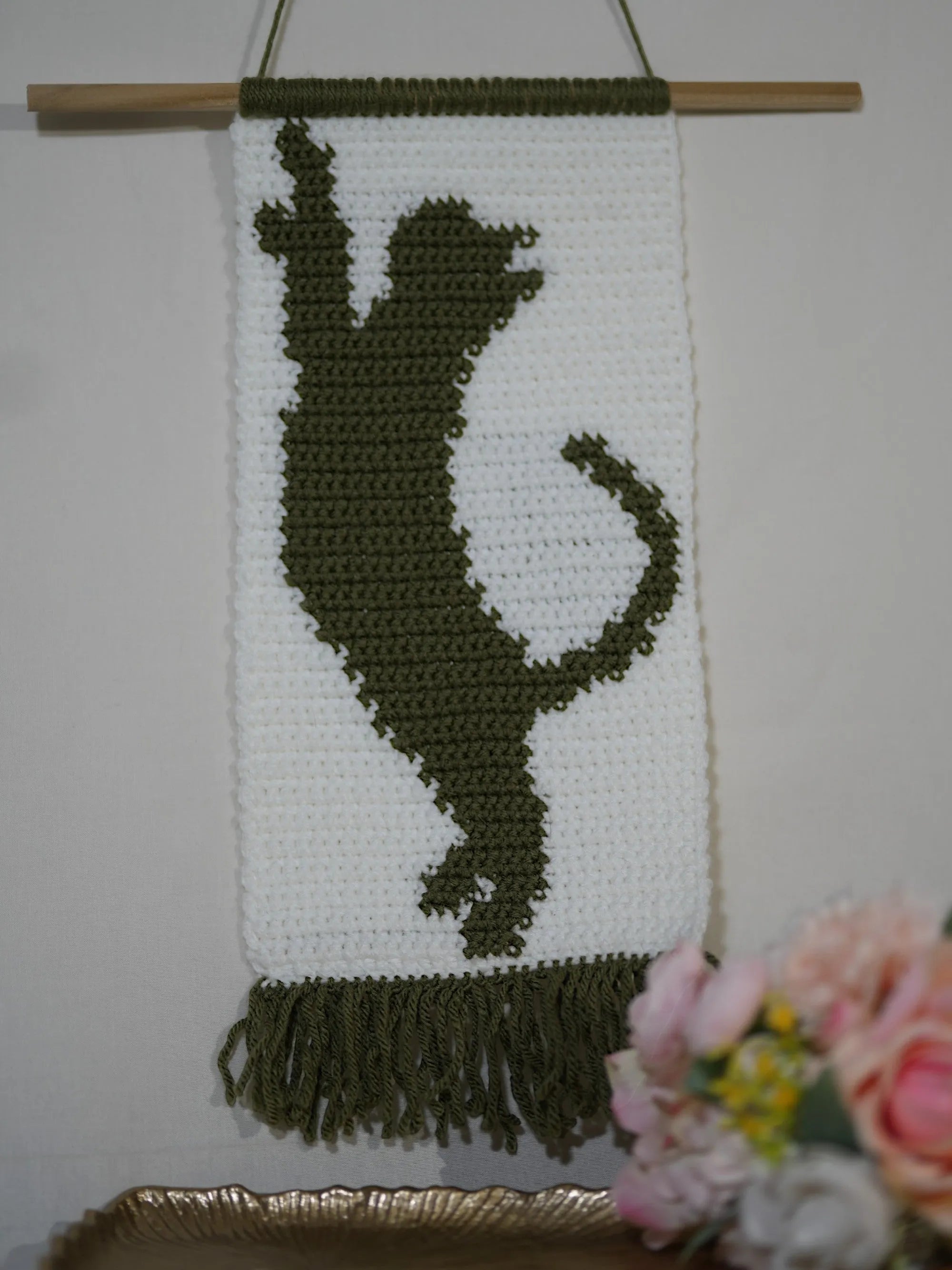 A woven wall hanging depicting a silhouette of a playful cat in dark yarn on a white background, featuring fringe at the bottom and mounted on a wooden dowel.