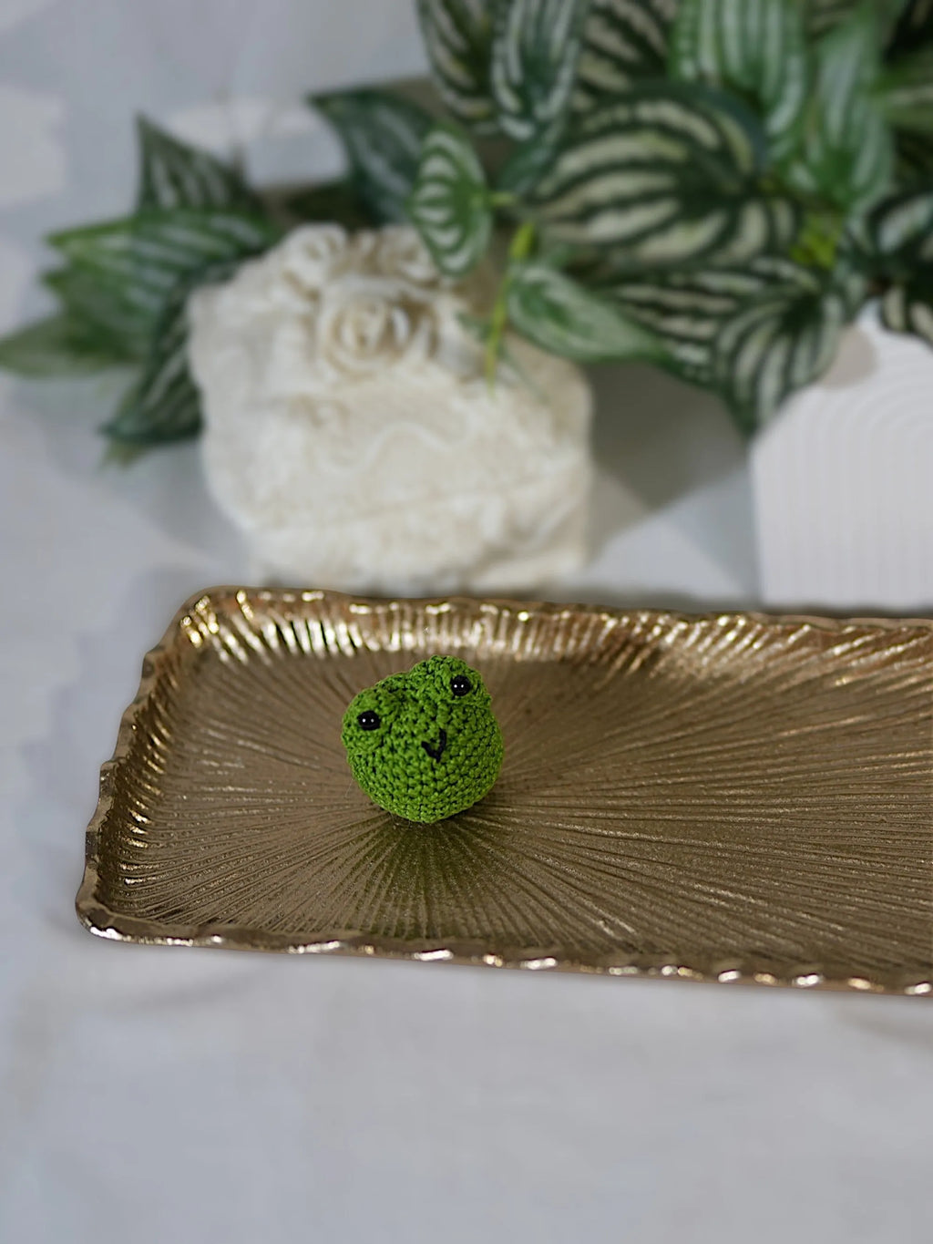 A small, crocheted green frog rests on a decorative gold tray with plants in the background.