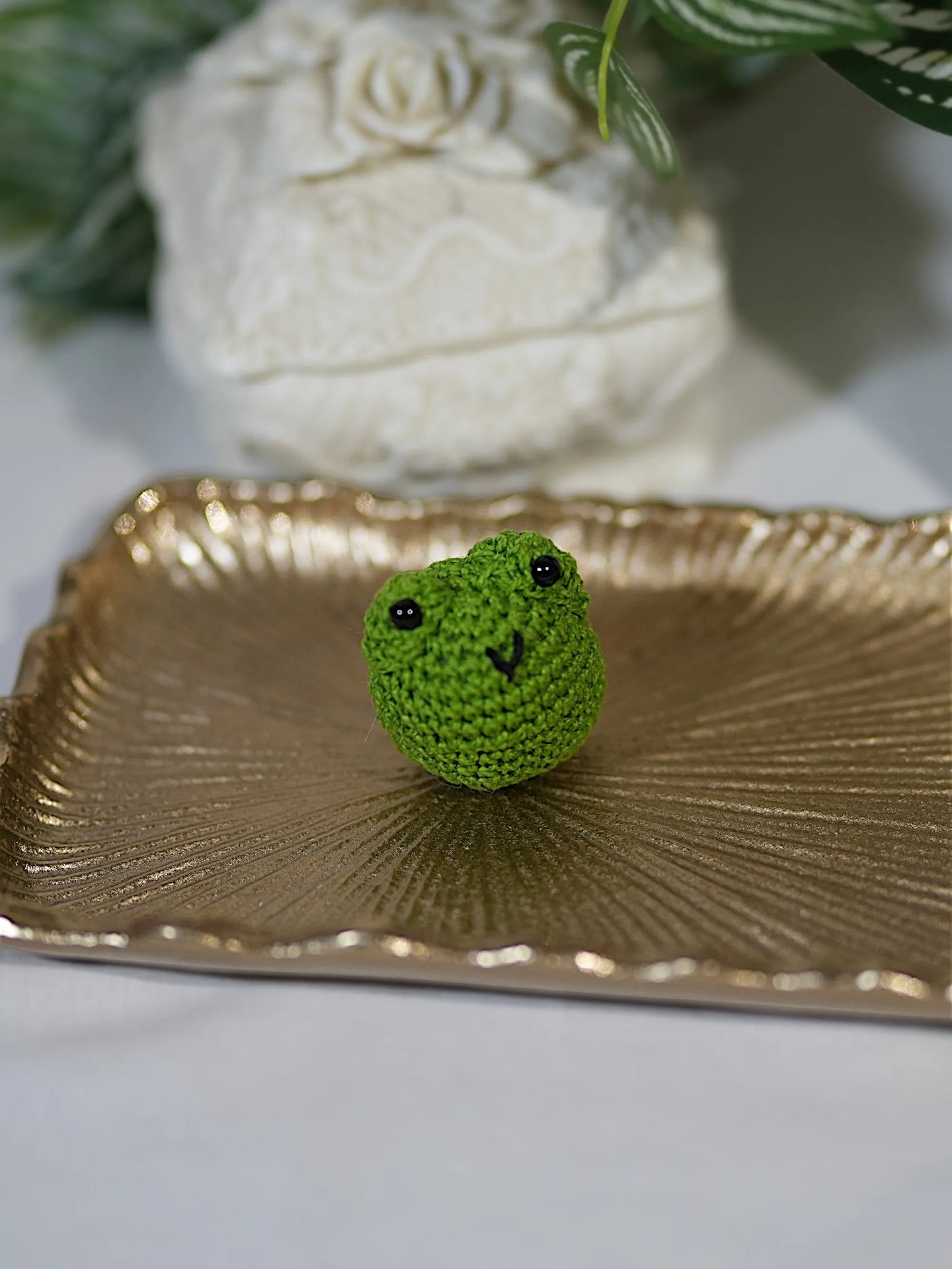 A small, hand-crocheted green frog with black eyes, sitting on a decorative golden tray with an ornate background.