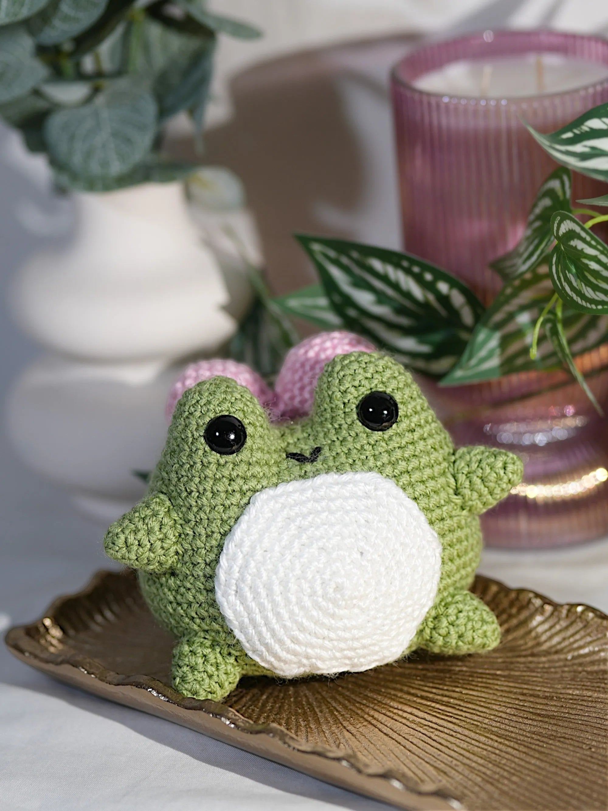 A cute green and white crocheted frog plush toy sitting on a decorative gold tray with leafy plants and a pink candle in the background.