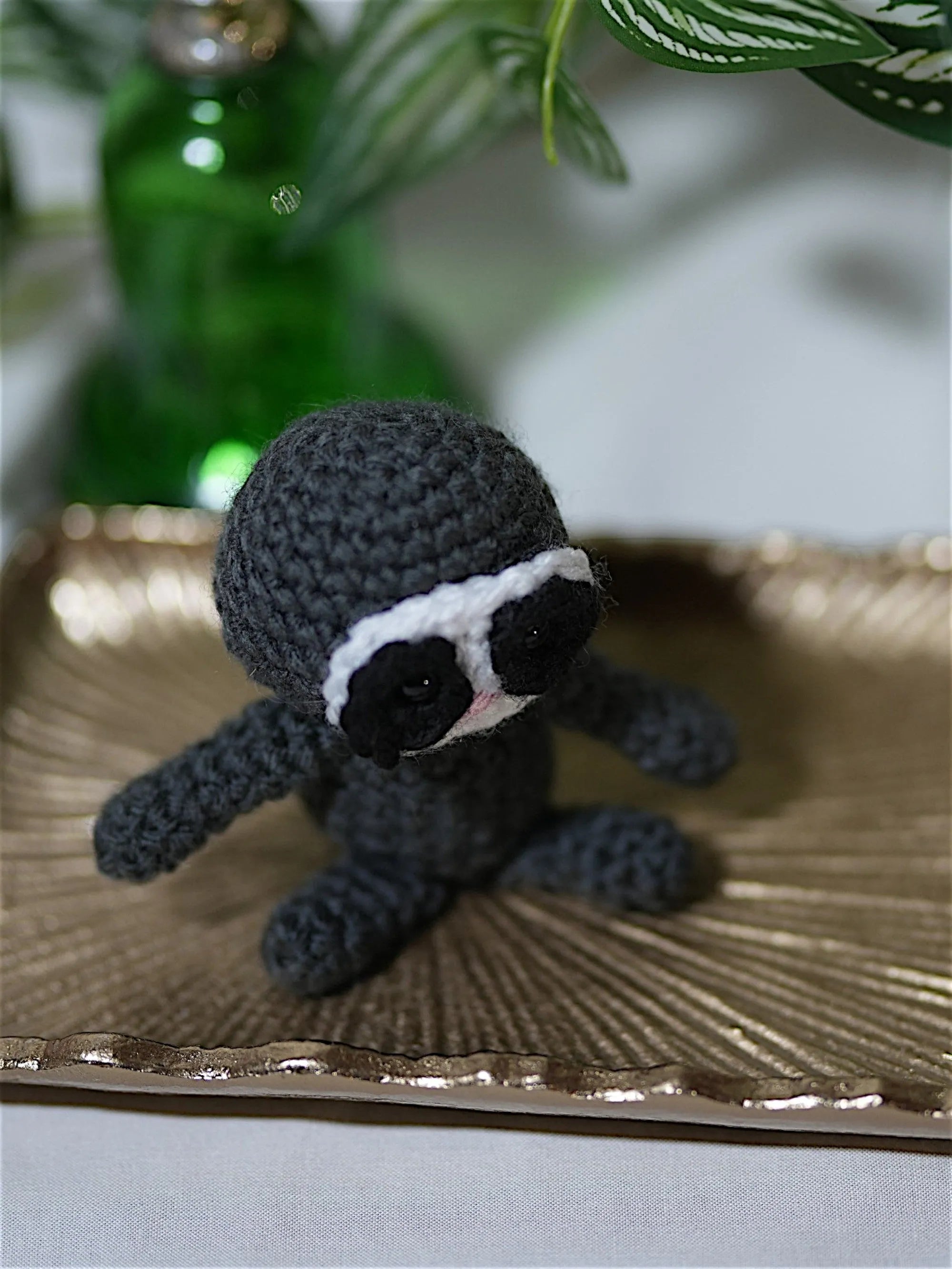 A small, crocheted gray sloth toy with black and white facial features, sitting on a decorative gold tray with green foliage in the background.