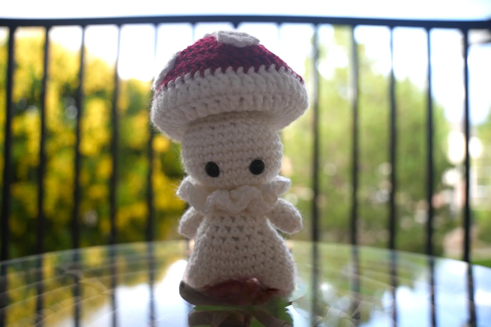 A crocheted mushroom character with a red and white cap, standing on a glass surface outdoors.