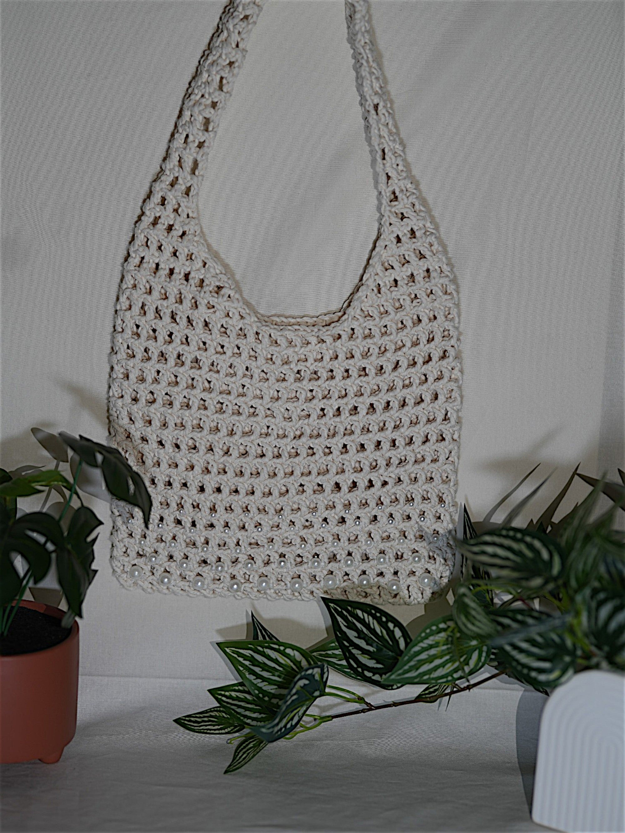 A handmade crocheted white bag hanging between two potted plants, featuring a textured pattern and pearl embellishments.