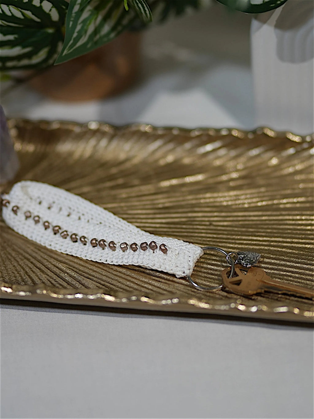 A stylish, handmade keychain with white crochet and bead details, resting on an elegant gold decorative tray.