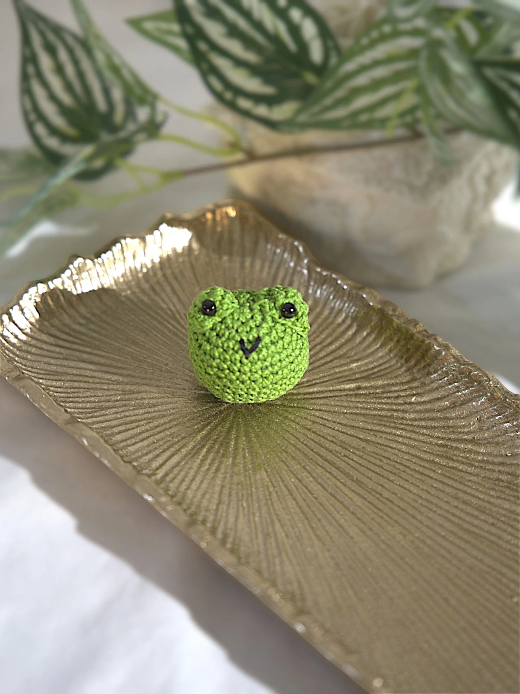 A small knitted green frog sits on a textured gold tray with decorative green plants in the background.
