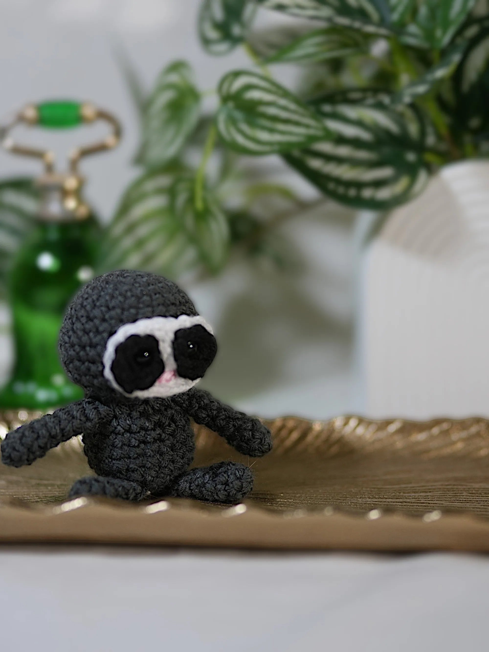 A cute knitted sloth figurine sitting on a decorative gold tray with green plants in the background.