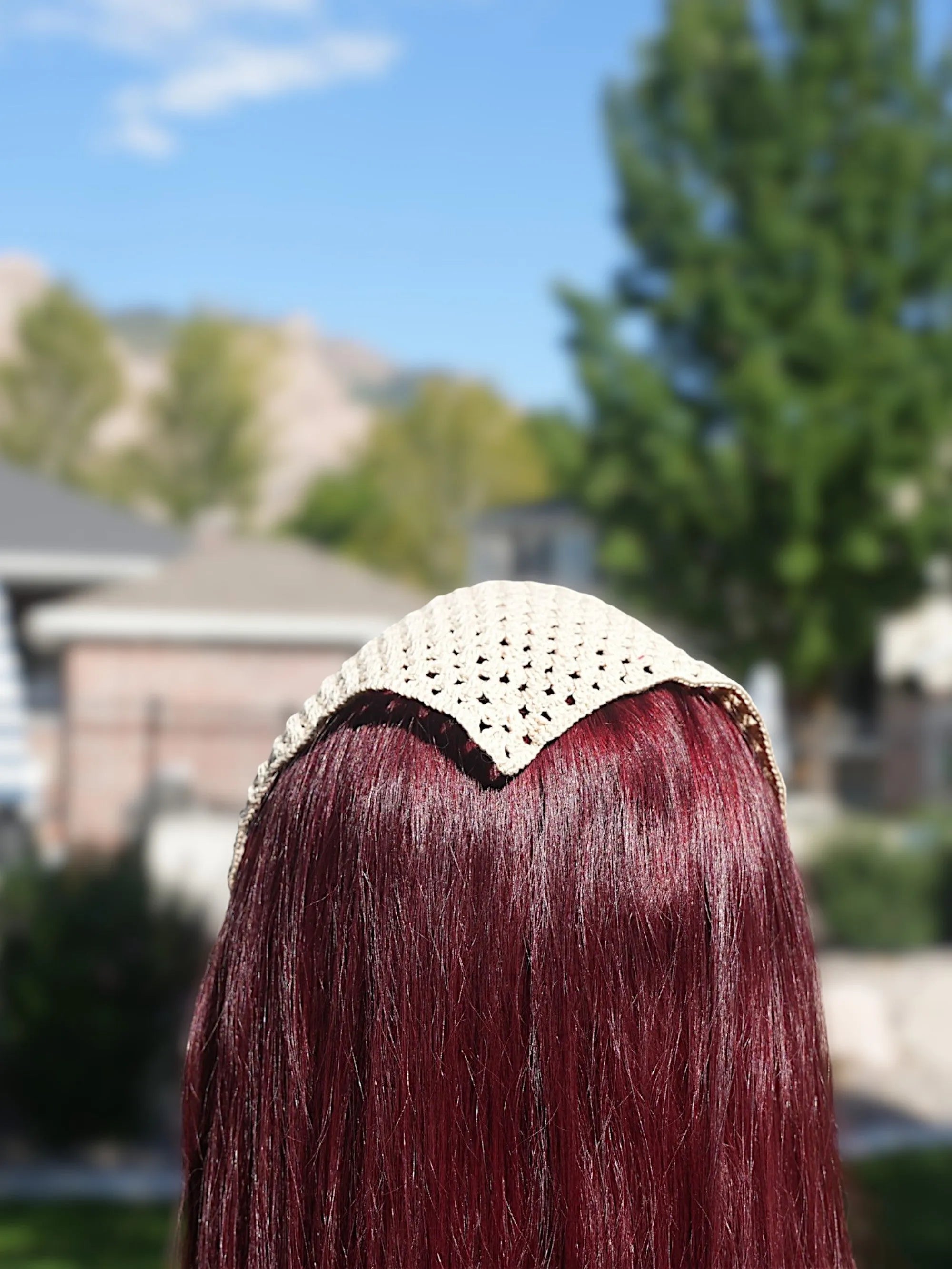 A woman with long red hair wearing a beige knitted headscarf standing outdoors.