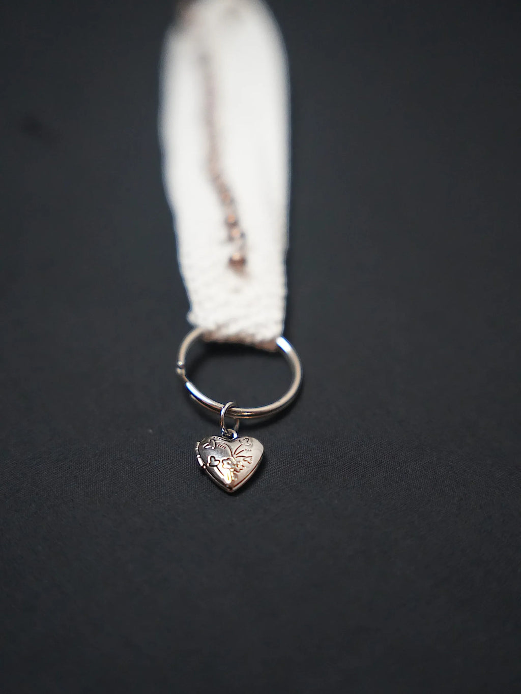 A silver heart-shaped charm with an engraved floral pattern attached to a metal ring on a cream-colored fabric strap.