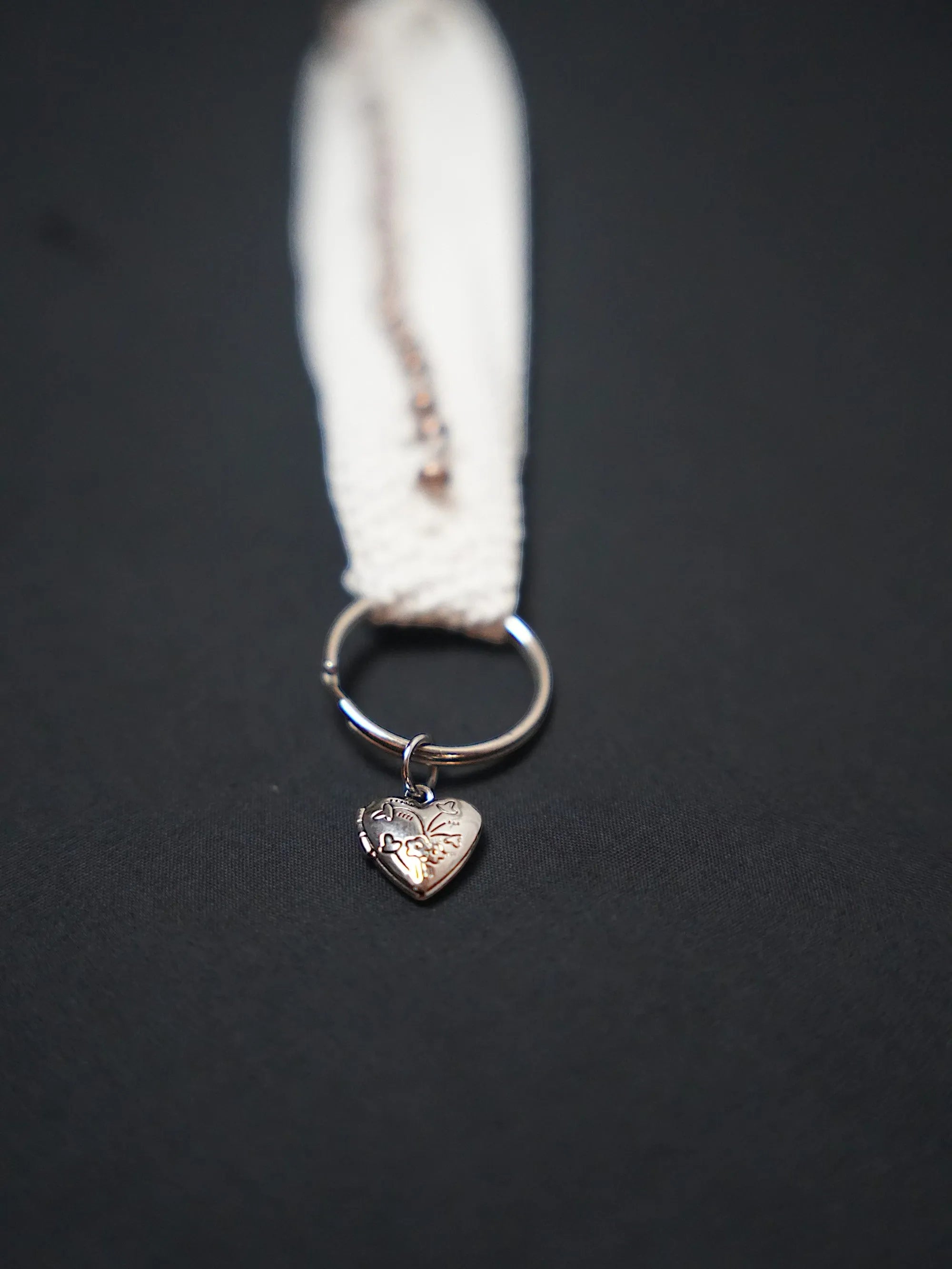 A silver heart-shaped charm with an engraved floral pattern attached to a metal ring on a cream-colored fabric strap.