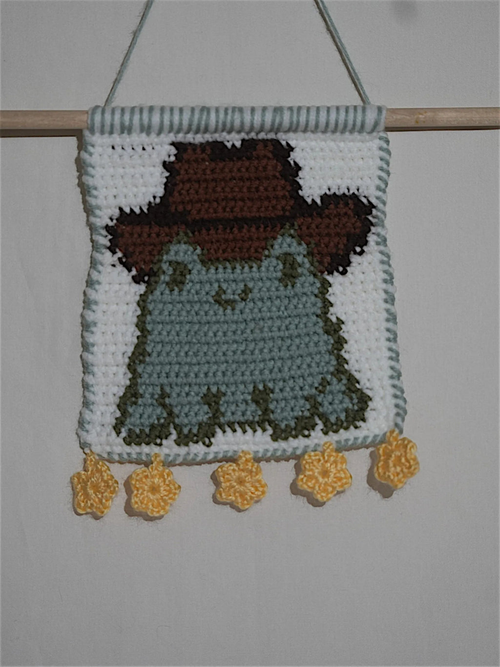 A crocheted wall hanging featuring a cute frog with a brown hat, embellished with yellow flower ornaments at the bottom.
