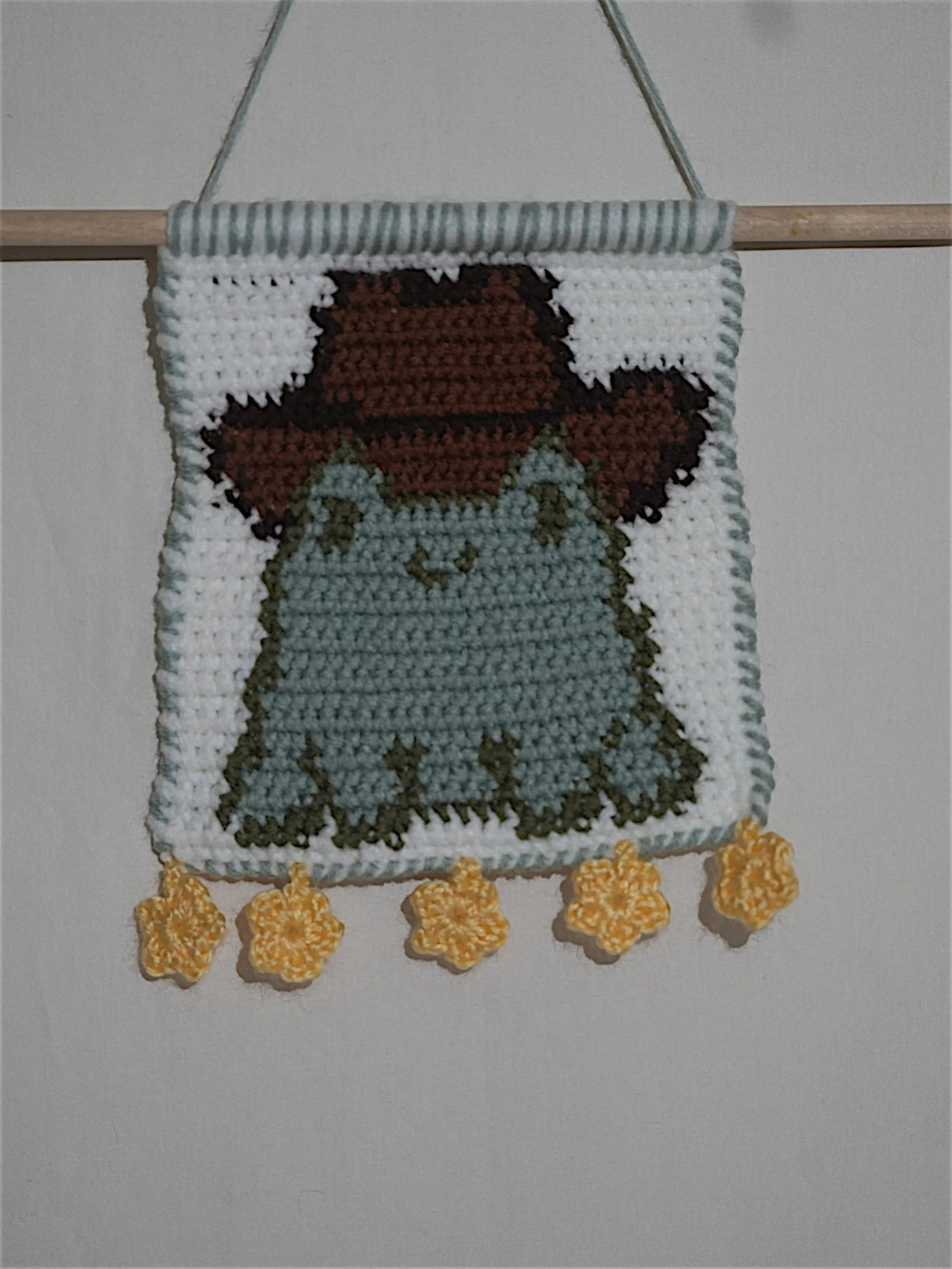 A crocheted wall hanging featuring a cute frog with a brown hat, embellished with yellow flower ornaments at the bottom.