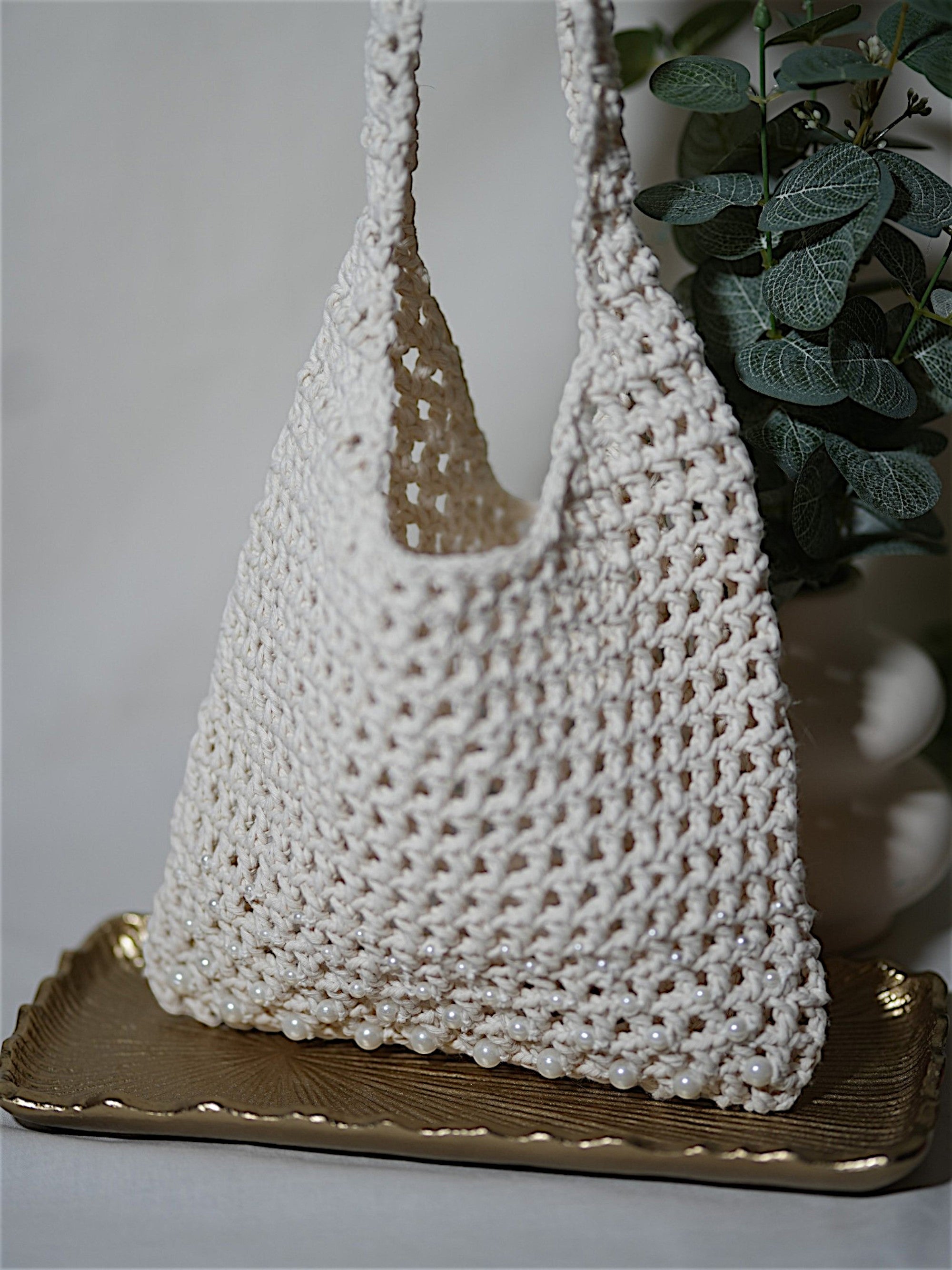 A handmade white crochet bag with pearl embellishments, displayed on a gold tray next to a green plant.