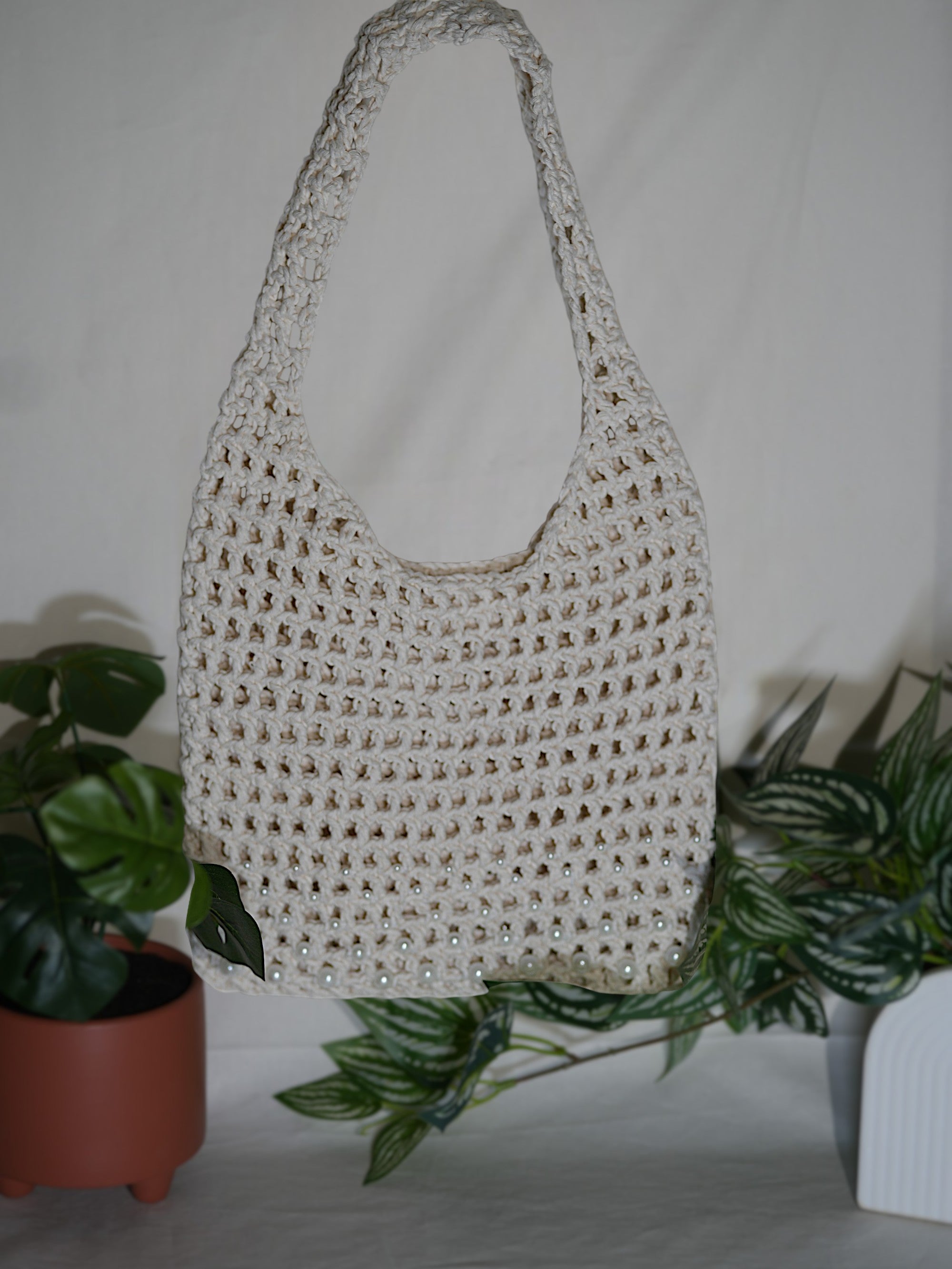 A handmade white crochet tote bag hangs in front of a backdrop with green plants and a terracotta pot.